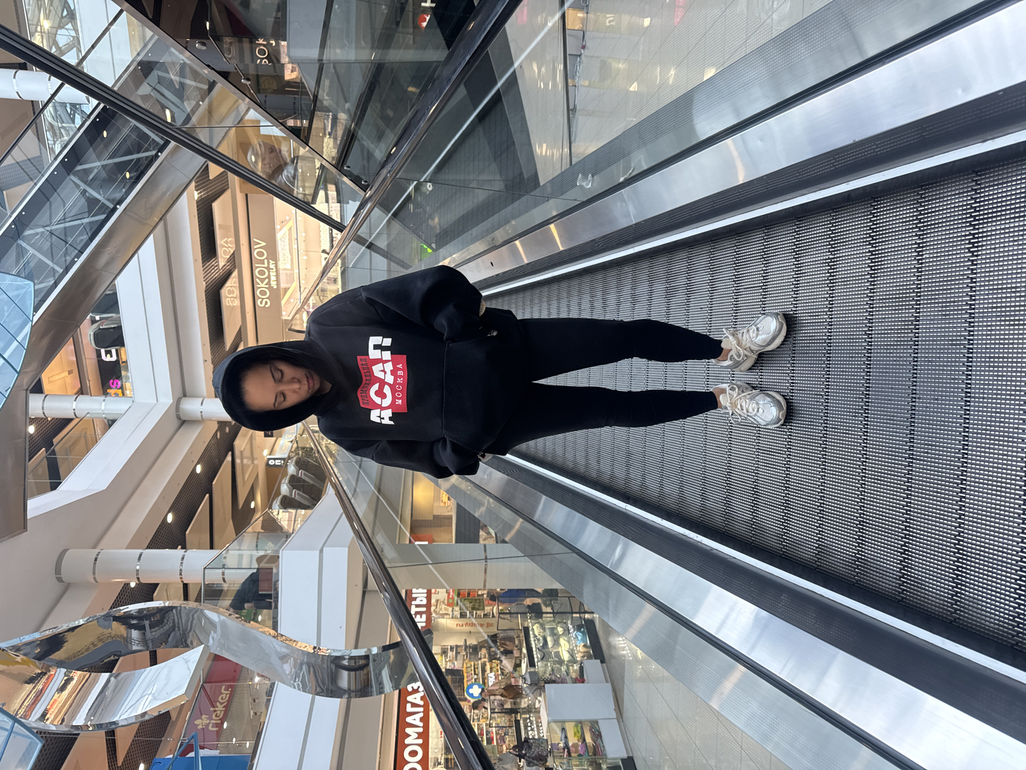 This media file shows a person standing on an escalator in a shopping mall. The person is wearing a black hoodie with red and white lettering that reads "ACAN МОСКВА" and black leggings. They are also wearing white athletic shoes. The escalator is moving upwards, and the background shows a multi-level shopping mall with various stores visible, including "SOKOLOV JEWELRY" and "rieker." The lighting suggests it is daytime. The person's head is tilted down, and their eyes are closed, giving an impression of relaxation or contemplation. The overall setting is a modern, well-lit indoor shopping environment. Text visible in the image includes store names and some Russian words.