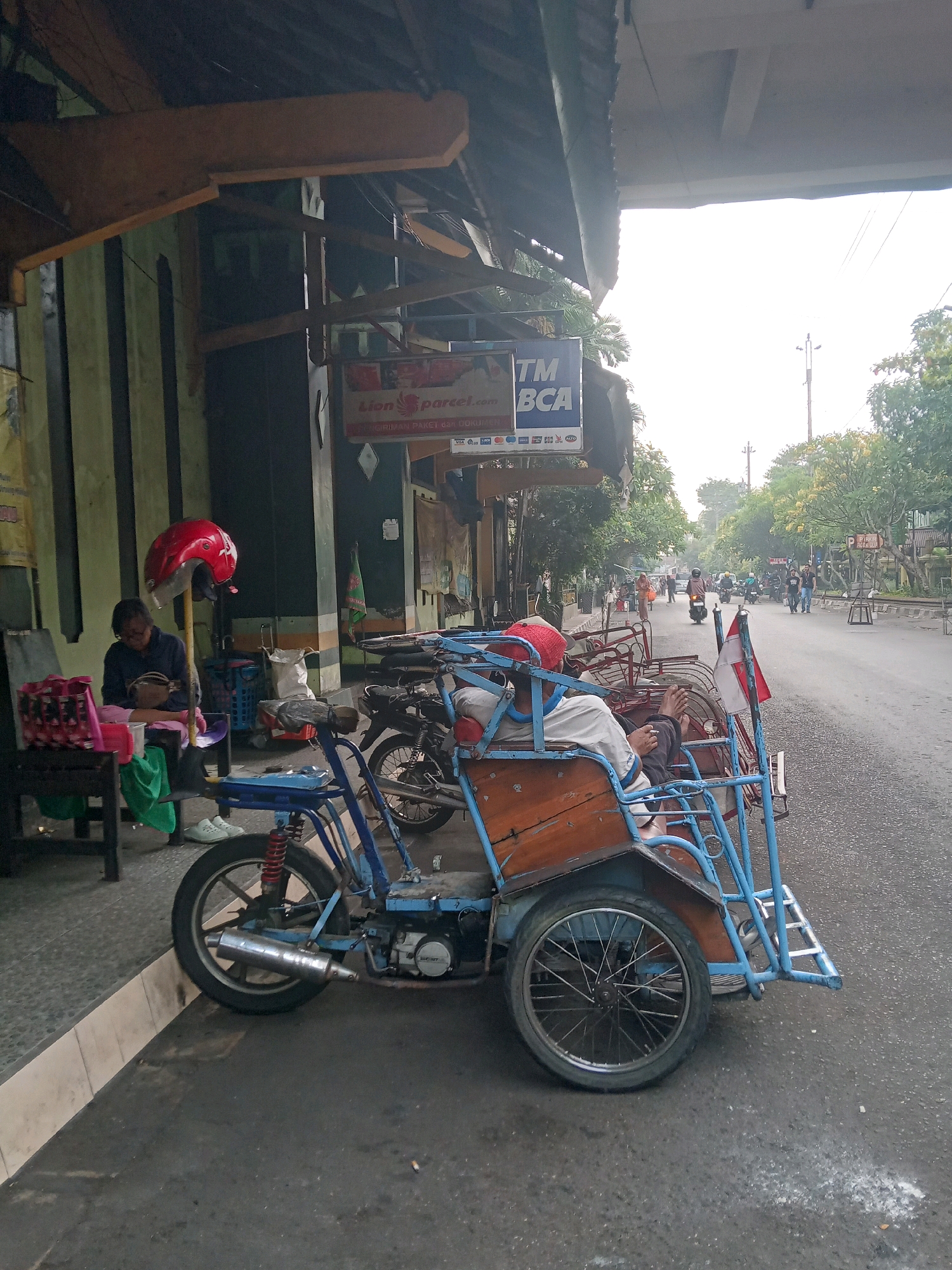 The image depicts a street scene in Yogyakarta, Indonesia. A blue and orange pedicab, a three-wheeled vehicle, is parked on the side of a paved road. The pedicab has a wooden passenger compartment with a blue metal frame. A person, wearing a red cap, white shirt, and dark pants, is seated in the pedicab, facing away from the camera, with their legs extended. A small Indonesian flag is attached to the pedicab's frame.
To the left of the pedicab, a person is seated on a chair near a building. This person is wearing glasses, a dark blue shirt, and a green cloth draped over their lap. A red helmet is placed on a stand beside them. In front of the seated person are bags and a small table.
The building behind them has a wooden overhang and green-striped walls. A sign for "TM BCA" and "Lion Parcel" is visible on the building. The road in front of the pedicab has other vehicles, including motorcycles, and several people walking. Trees line the other side of the road, and the sky is overcast. The ground appears wet, suggesting recent rain.