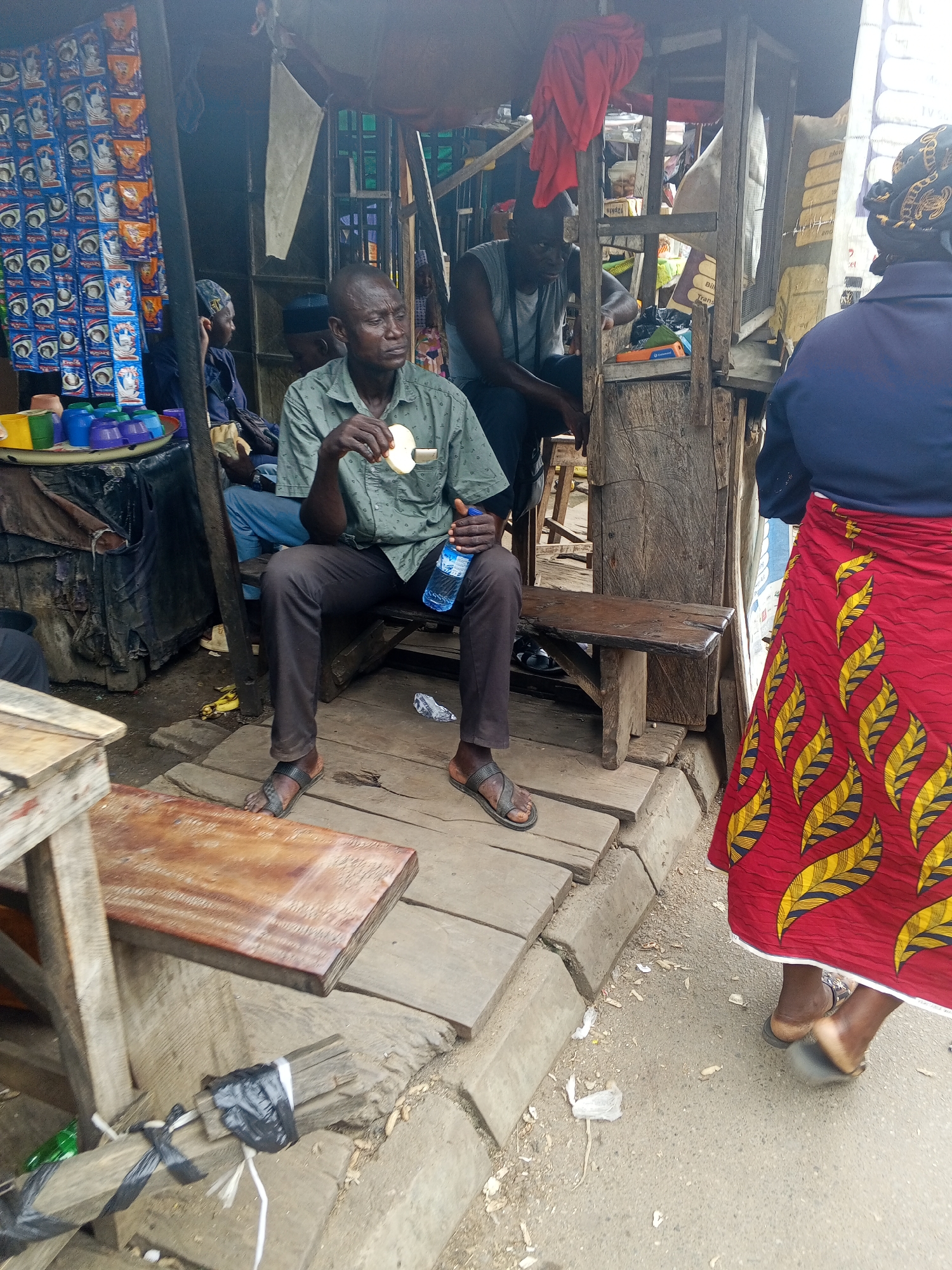 The image depicts a street scene, likely in Okene, Nigeria, with an outdoor market stall. A man in a green, short-sleeved, collared shirt and dark pants sits on a wooden bench, holding a slice of food and a bottle of water. He is wearing sandals. To his right, another man in a grey tank top and dark pants also sits, looking towards the front. Further in the background, a person wearing a blue top and a red skirt with a yellow leaf pattern stands with their back to the camera. The market stall features a wooden structure, with shelves displaying various items, including colorful plastic cups on the left. The ground is made of wooden planks and concrete. The overall environment appears to be that of a bustling local marketplace.