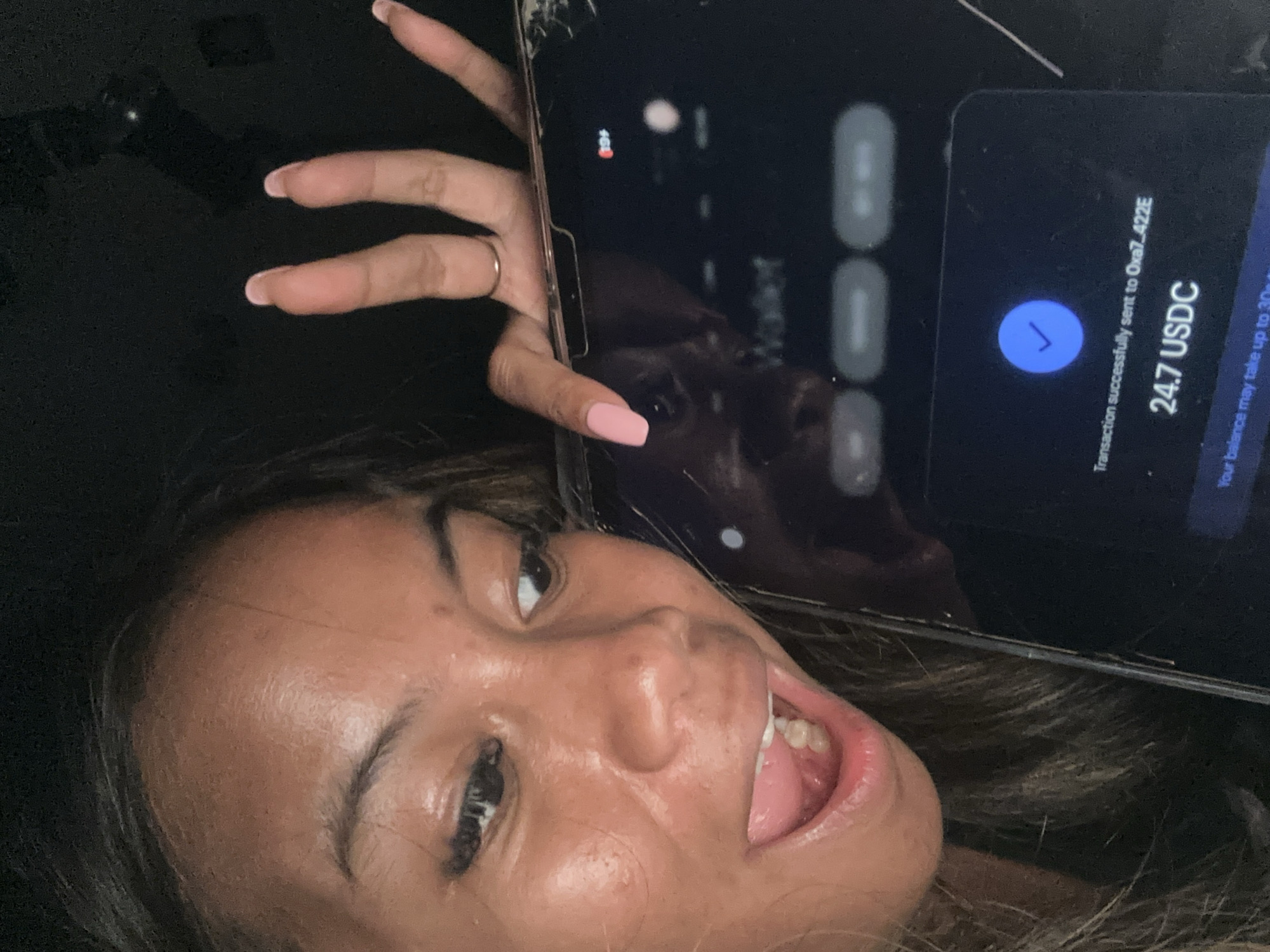 A person is holding a cracked phone displaying a successful transaction of 24.7 USDC to 0xa7_422E. The background appears dark. The person is facing the camera with an open mouth and partially extended tongue. No information within this image reveals the location of the city of North Jakarta, Indonesia.