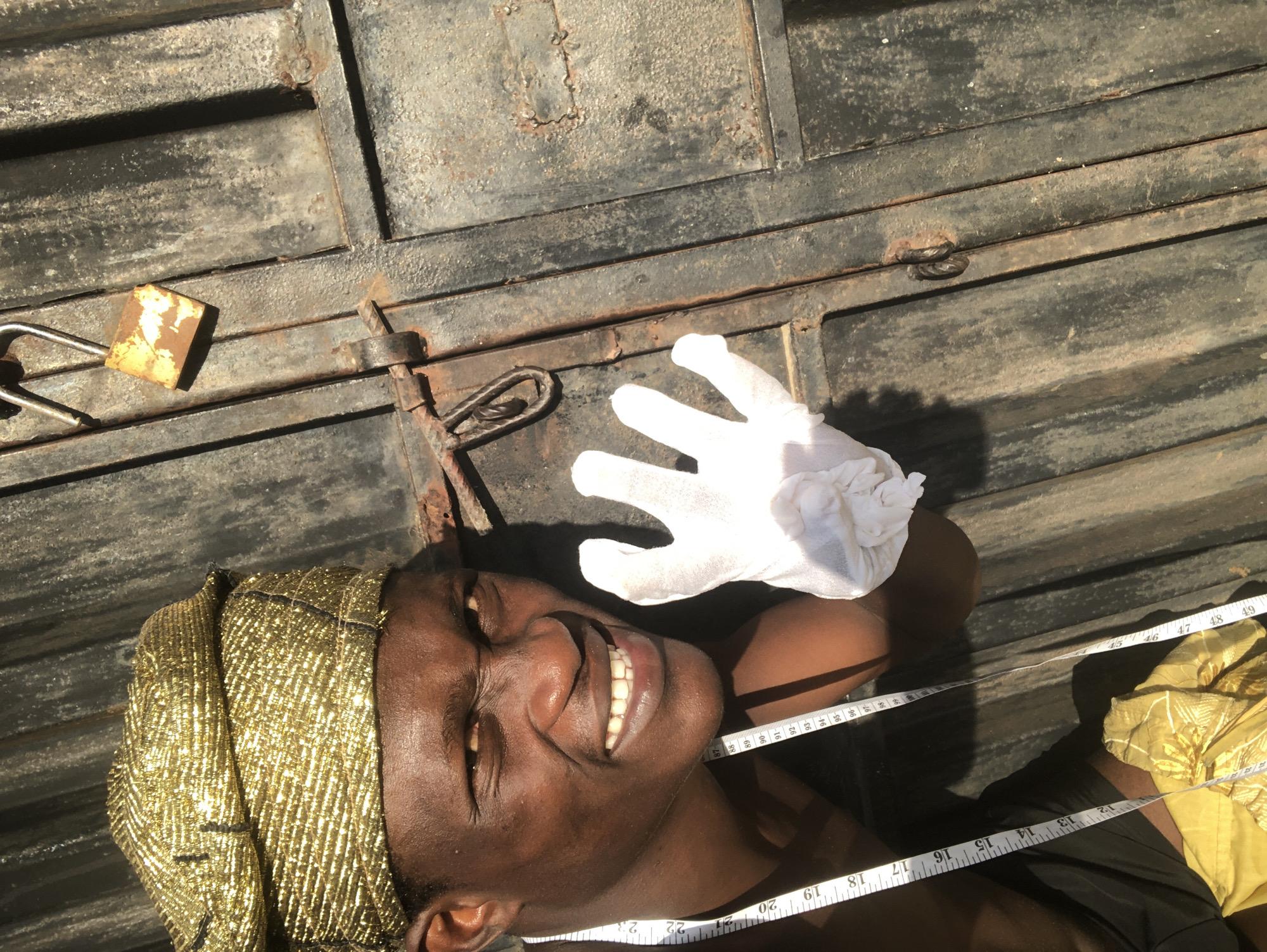 An outdoor, eye-level, close-up shot captures a Black man wearing a gold cloth headwrap and a white glove on his right hand. He is leaning towards a rusty metal door and making a facial expression. The man is also wearing a white tape measure around his neck, with markings visible down his torso. To the left of the man is a yellow cloth with gold embellishments. The door, which appears to be made of corrugated metal, has a rusty latch and a small, rusty padlock attached to it. The lighting suggests it is daytime, with shadows cast on the door, indicating the sun is to the left of the frame.