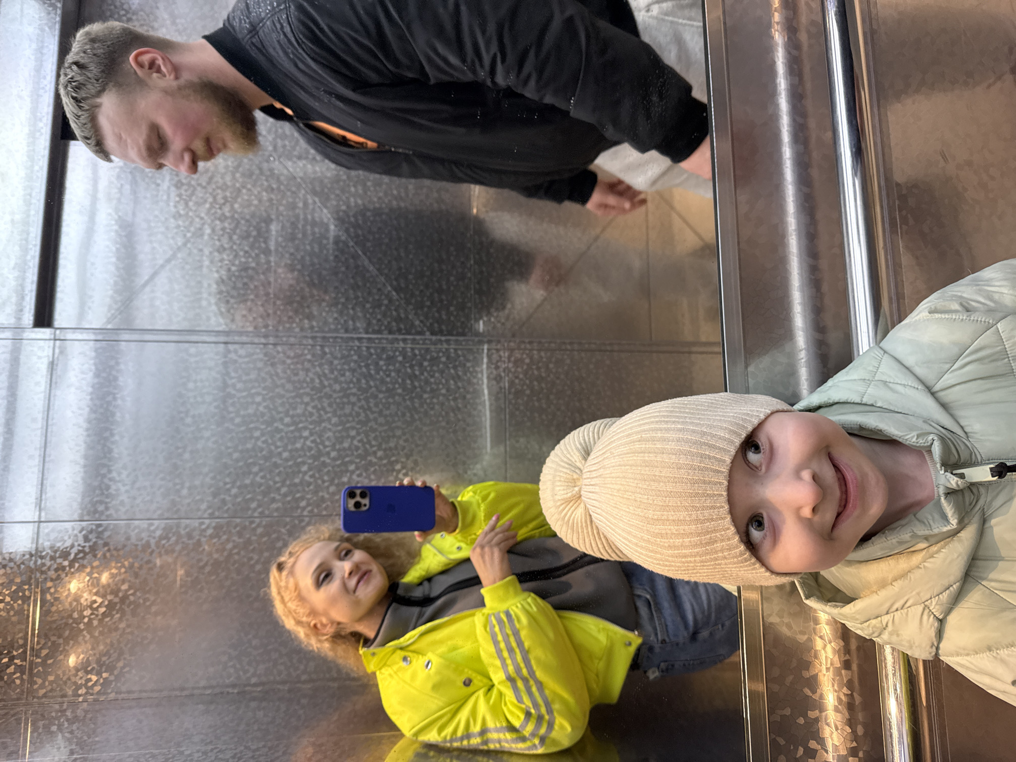 In Perm, Russia, two women and a man are in an elevator with mirrored walls. One woman in a yellow jacket takes a selfie with a blue cell phone. The other woman wears a beige beanie and a green jacket. The man wears a black jacket. They are reflected in the elevator's mirrored surfaces.