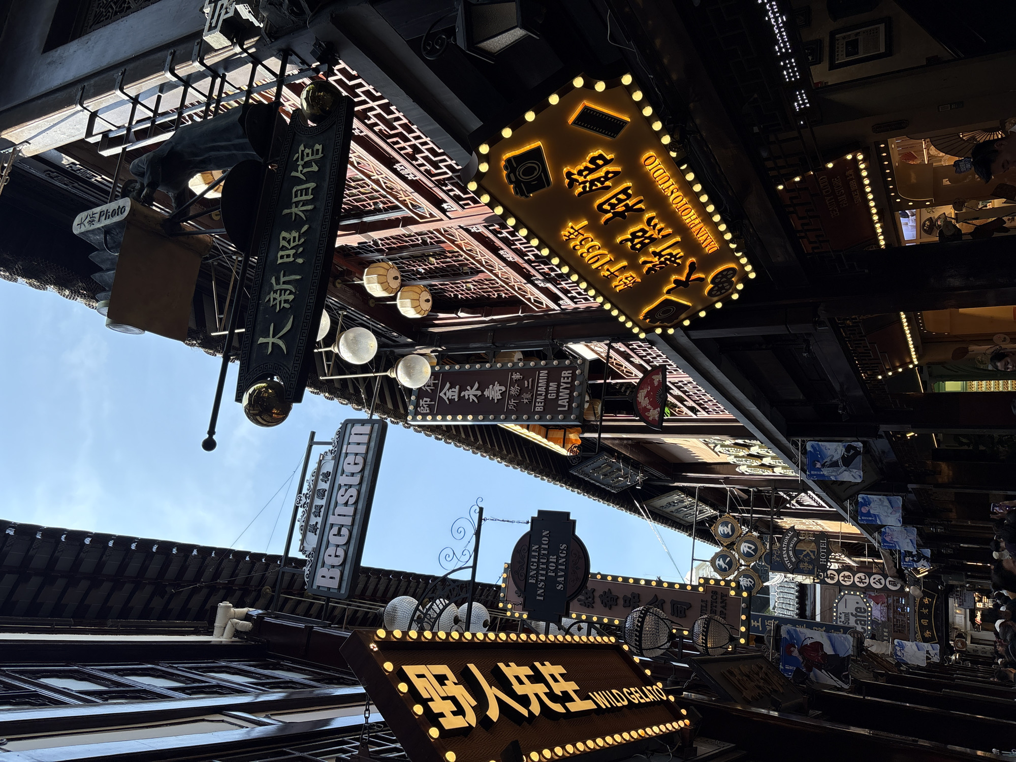 This is a low-angle, eye-level shot of a bustling street in Shanghai, China, likely the Yu Garden Bazaar, known for its traditional architecture and lively atmosphere. The buildings on either side of the narrow street are several stories high and feature dark, ornate wooden facades with intricate lattice work on the upper levels. A bright blue sky with a few wispy clouds is visible overhead, peeking through the narrow gap between the buildings.
The street is lined with numerous signs, many of them illuminated with bright yellow lights, indicating various businesses. Prominently displayed is a large, neon-yellow sign with Chinese characters and the English text "DAXIN PHOTO STUDIO," featuring graphics of a camera and film roll. Another sign, also illuminated, reads "WILD GELATO" in large, bold font. Several other signs in Chinese are also visible, some with accompanying graphics of people or objects. Traditional lanterns and decorative light fixtures hang from the buildings, adding to the festive ambiance.
There are also some notable English signs, including one that says "Bechstein," likely for a piano store, and another that reads "ENGLIN INSTITUTION FOR SAVINGS." A sign for "HOTEL" is also visible further down the street. Hanging banners with illustrations of anime-style characters are displayed on some of the buildings.
The street is populated by a crowd of people, most of whom are not clearly discernible but appear to be tourists and locals strolling and browsing. Some individuals are looking at their phones, while others are engaged in conversation or looking at the shops. The overall impression is one of a vibrant, commercial area filled with a mix of traditional and modern elements. The sunlight casts strong shadows, highlighting the depth and texture of the scene.