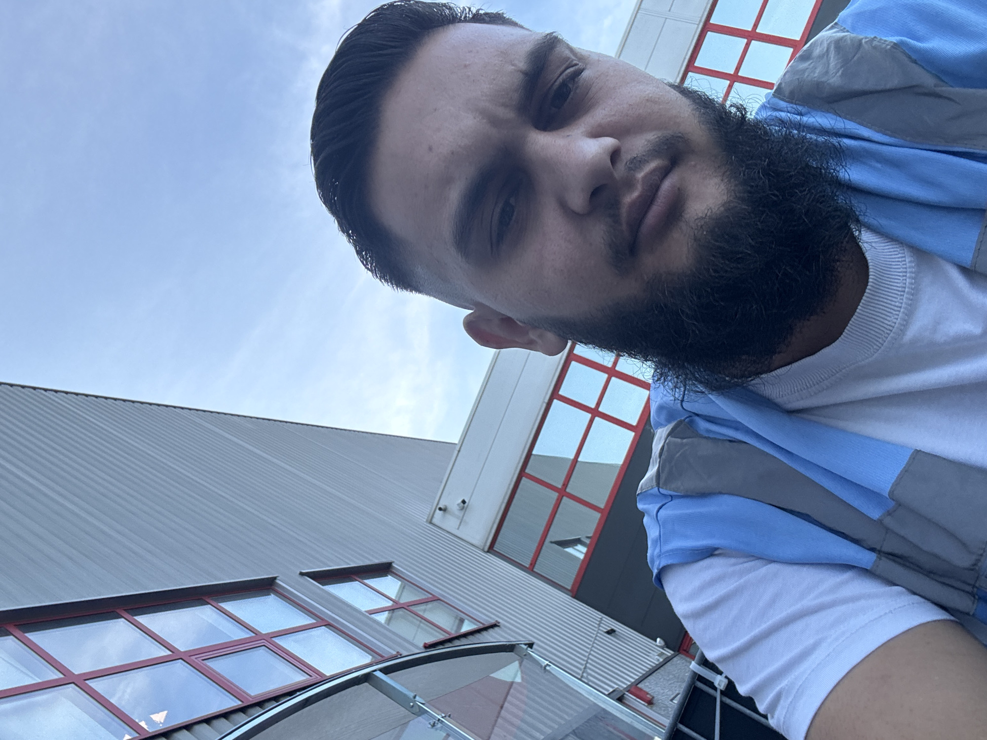 A low-angle selfie is taken outdoors, with the subject's face dominating the foreground. The man has dark hair, a full beard, and is wearing a white t-shirt layered with a blue and grey vest. In the background, a modern building with ribbed metal siding and large windows with red framing is visible. The sky above is clear and blue. The location is the city of Boom, Belgium.