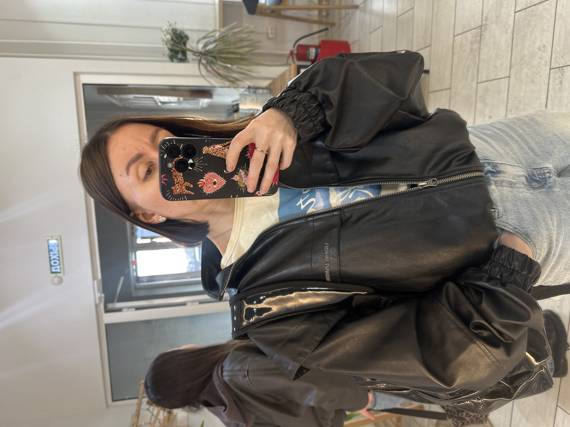The image depicts an indoor scene within a commercial or public establishment in Votkinsk, Russia, as evidenced by a reversed green "ВЫХОД" (EXIT) sign above a glass door in the background.
A woman is positioned centrally, taking a mirror selfie with a dark-colored smartphone featuring a distinctive case with leopard print and sacred heart motifs. Her face is partially obscured by the phone. She has brown hair, wears a light-colored t-shirt with blue text, a black bomber-style jacket with a zippered front and elasticized cuffs, and light blue denim jeans. A glossy black strap, possibly from a backpack, is visible over her left shoulder. She wears a gold-colored ring on her left ring finger and a heart-shaped earring on her visible ear. Her left hand is tucked into her jacket pocket.
In the background reflection, another individual, a woman with dark brown hair, is partially visible from behind, wearing a brown hooded garment.
The environment features light-colored walls and a light-toned tiled floor. To the right of the primary subject, a potted plant stands, and further back, a red fire extinguisher is visible on the floor. Elements such as a wooden bench or counter and a dark bar stool are also discernible in the reflections. The visible text "USHU EHSU" (mirrored) appears on the front of the primary subject's jacket.