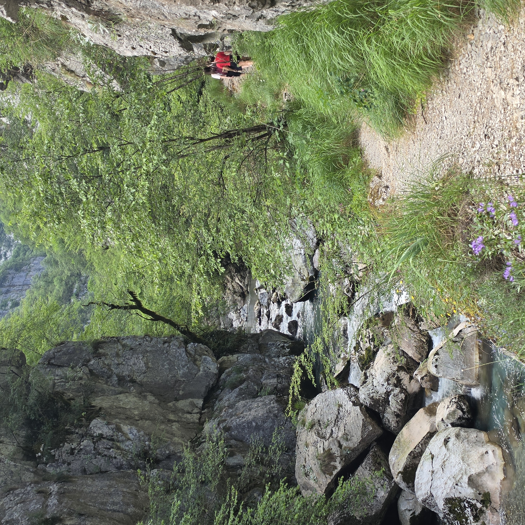 The image depicts a rocky gorge with a stream running through it in Lazba, Montenegro. The streambed is filled with large, light-colored rocks. Lush green trees and vegetation grow along the banks and on the rocky cliffs. A narrow gravel path runs along the upper right side of the image. Two figures wearing red shirts are walking along the path in the background.