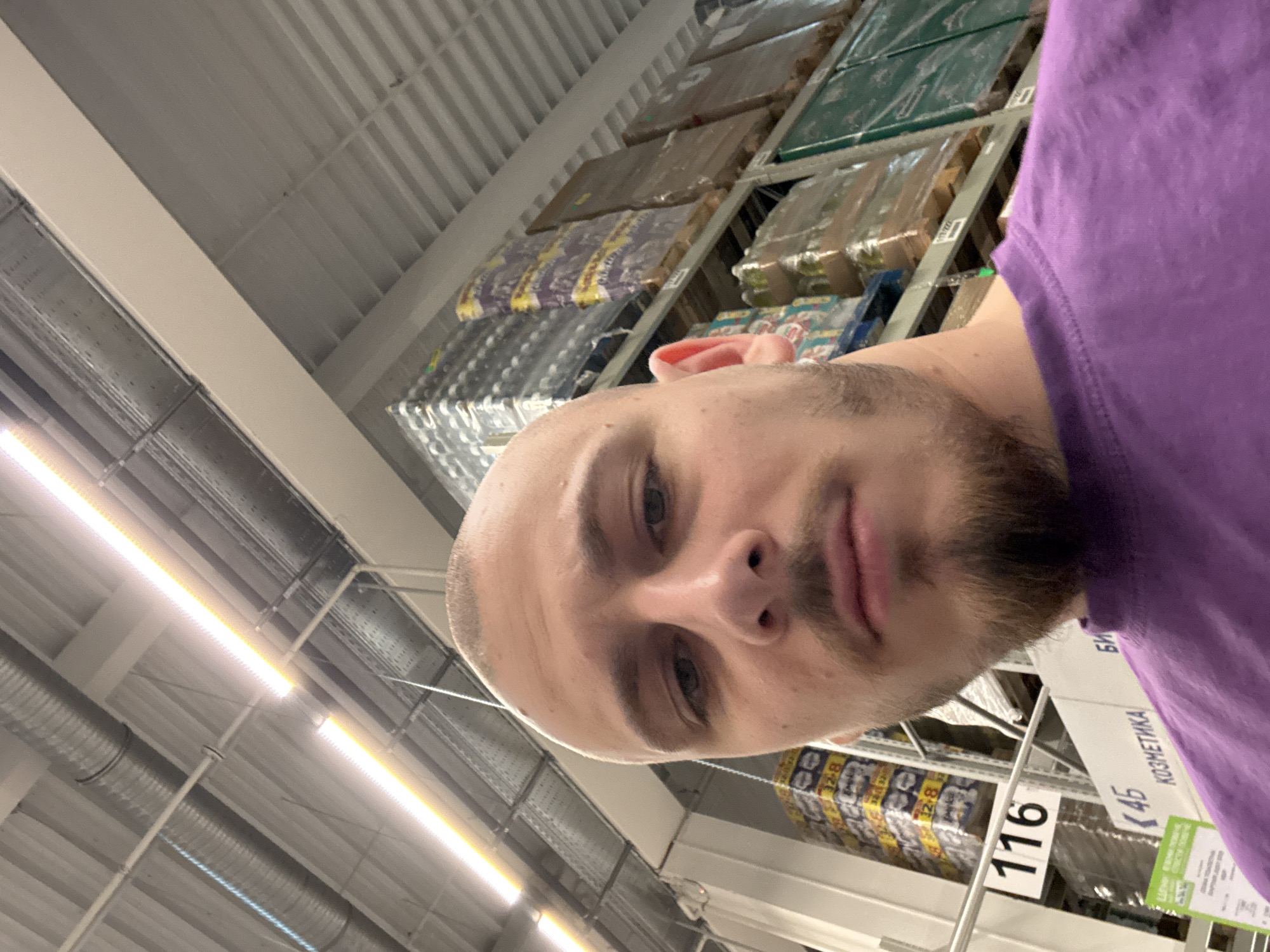 The image depicts a man taking a selfie in what appears to be a warehouse or large retail store. The man has a shaved head, facial hair, and is wearing a purple shirt. In the background, there are shelves stocked with various packaged goods and cosmetics products, along with product labels in Bulgarian that indicate the location is likely in Bulgaria. The sign visible in the photograph indicates that the subject is standing in front of cosmetics shelves. There is no definitive evidence to suggest that this specific location is in Stara Zagora, Bulgaria.