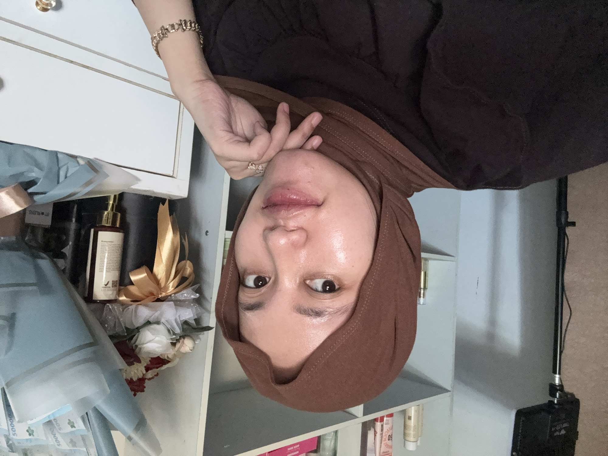 A woman wearing a brown hijab poses for a photo in an indoor setting. She is in front of a white shelving unit containing various items, including a bouquet of flowers and cosmetic bottles. The location may be Gadingrejo, Indonesia.