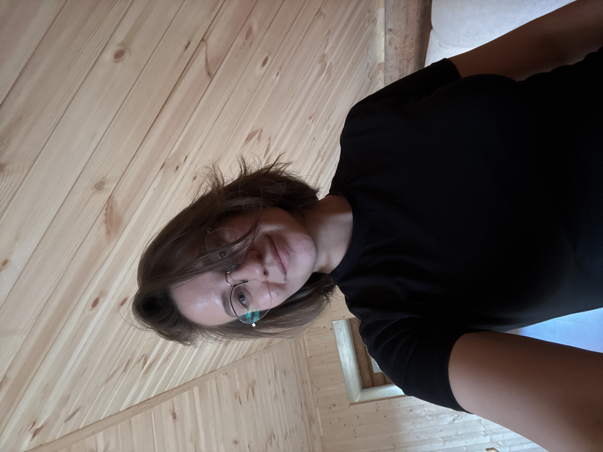 The image displays a female subject in an indoor setting within the city of Lenino, Russia. The subject is positioned centrally, viewed from the chest up, and appears to be taking a selfie, looking directly at the camera with a slight smile. She has short, dark brown hair, slightly tousled, and is wearing wire-rimmed glasses with lenses that exhibit blue-green reflections. Her skin is fair, showing some sheen on the forehead. She is dressed in a plain black short-sleeved t-shirt. The background features light-toned wooden plank paneling on both the walls and a distinctively sloped ceiling, suggesting an attic or room with a pitched roof. The wood exhibits prominent grain patterns and knots. To the left of the subject, a rectangular wooden-framed opening or doorway is visible. To the right, a segment of white, possibly upholstered, furniture or cushioning is present at the bottom right of the frame.