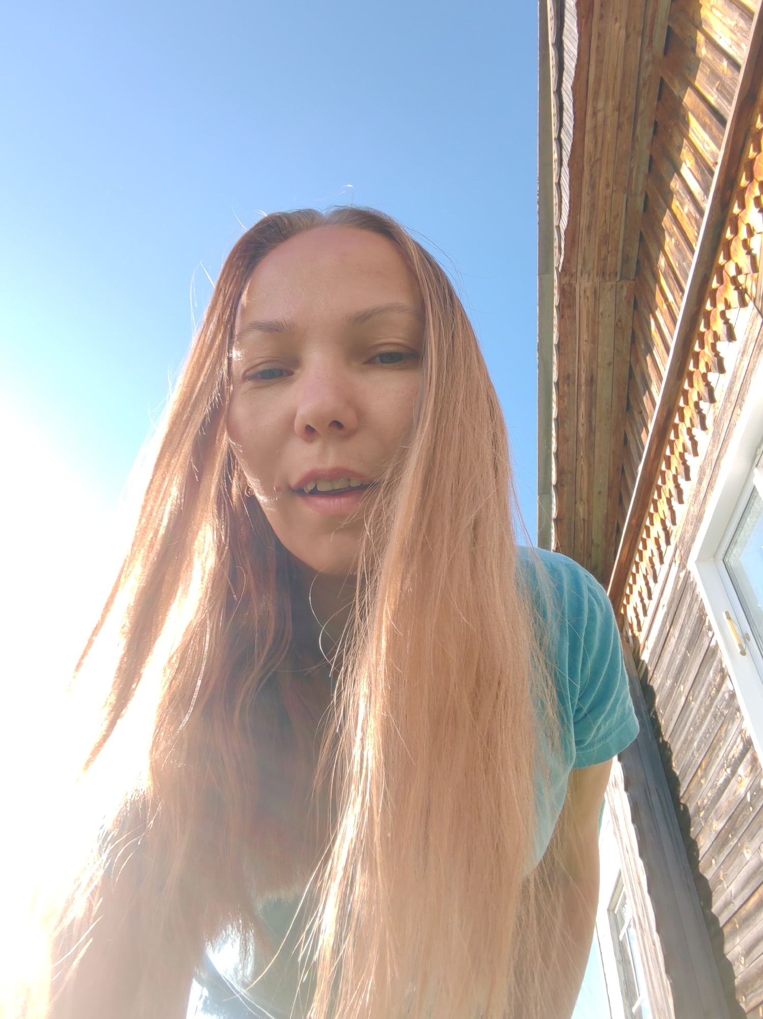 The image shows a young woman with long, light brown hair, wearing a teal t-shirt. She is looking down at the camera, with her face partially obscured by her hair. The background consists of a clear blue sky and a wooden building with intricate carvings. The light source appears to be the sun, positioned to the left of the frame, creating a lens flare effect. The setting seems to be outdoors, possibly in a rural or historic area, with the building suggesting a traditional Russian architectural style. The image was taken from a low angle, looking up at the woman.