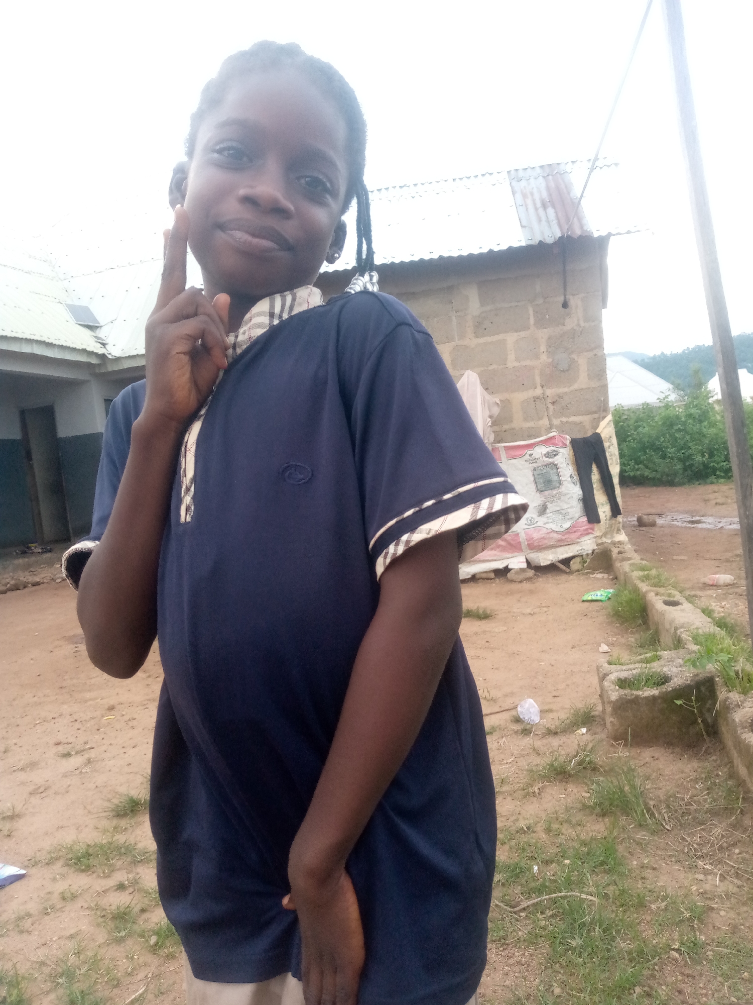 A young Black female stands in a semi-profile pose, facing towards the left. She has her right index finger raised towards her temple, with a slight smile on her face. She is wearing a dark blue t-shirt with a patterned collar. The background consists of a simple, rural setting with brick buildings, a corrugated metal roof, and dirt ground with sparse vegetation. The image appears to be taken outdoors, with overcast lighting. The location is identified as Serti, Nigeria.