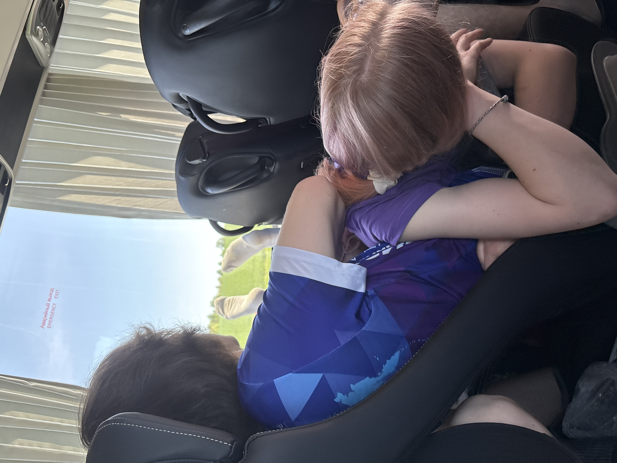 Two people are visible within the interior of what appears to be a vehicle, possibly a bus or van, based on the window and seat configurations. One person, with light brown hair, is seated and leaning over towards the other person. The seated person is wearing a purple shirt and blue shorts with white trim. The other person has dark brown hair and is reclined against a seat. A sign in Russian is visible on the window, translated to "Emergency Exit." The environment visible outside the window suggests a rural or suburban setting with trees and greenery. No information about the specific location in Semenovo, Russia can be determined from the image.