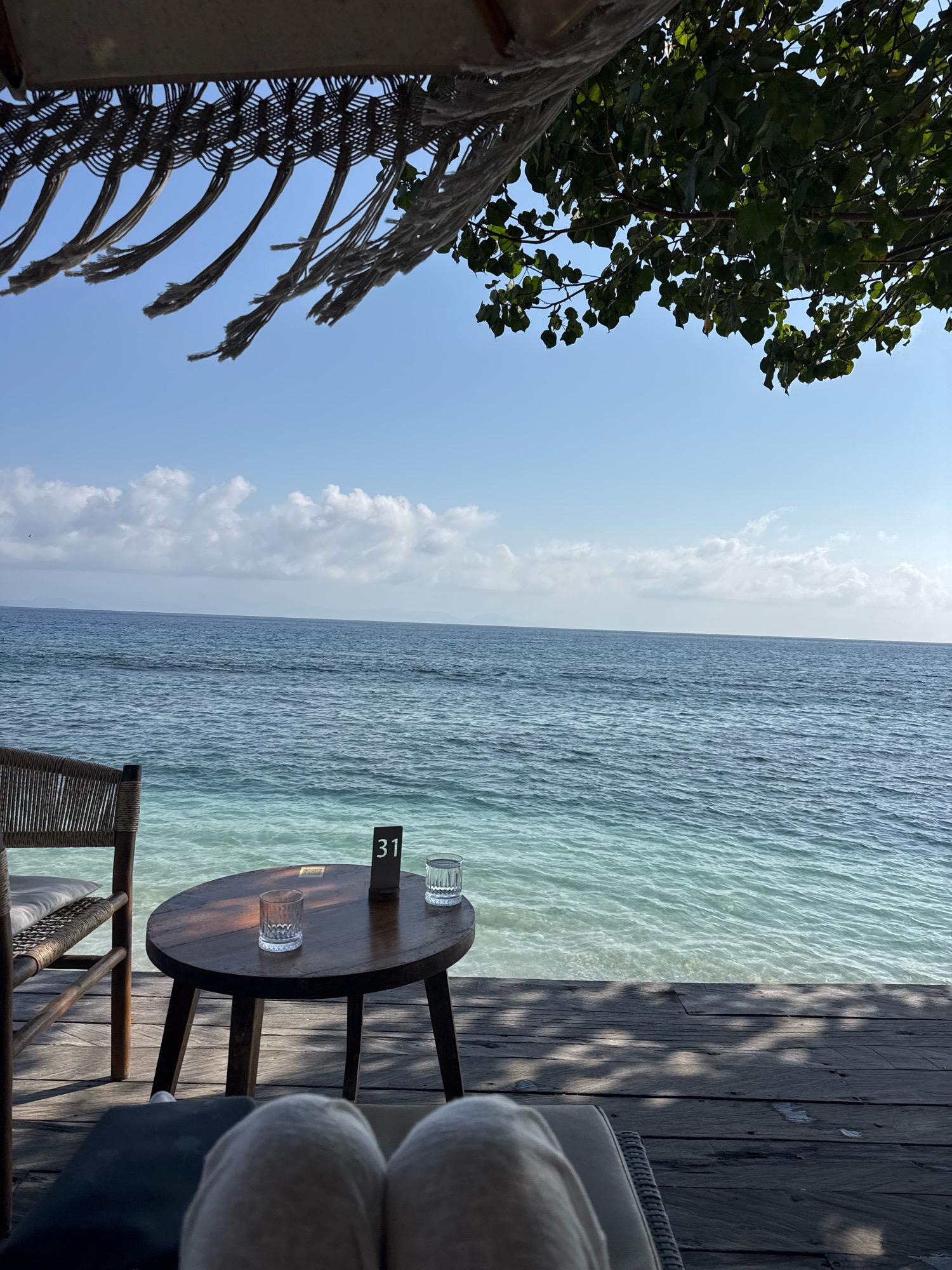 An outdoor perspective shows a wooden table with two glasses and a table number "31" on it. A chair is situated to the left of the table. The scene is set on a wooden deck with a view of the ocean. Above, a canopy with hanging ropes and leaves from a tree are visible, with a blue sky in the background. The lower portion of the image shows the legs of a person lying on a lounge chair.