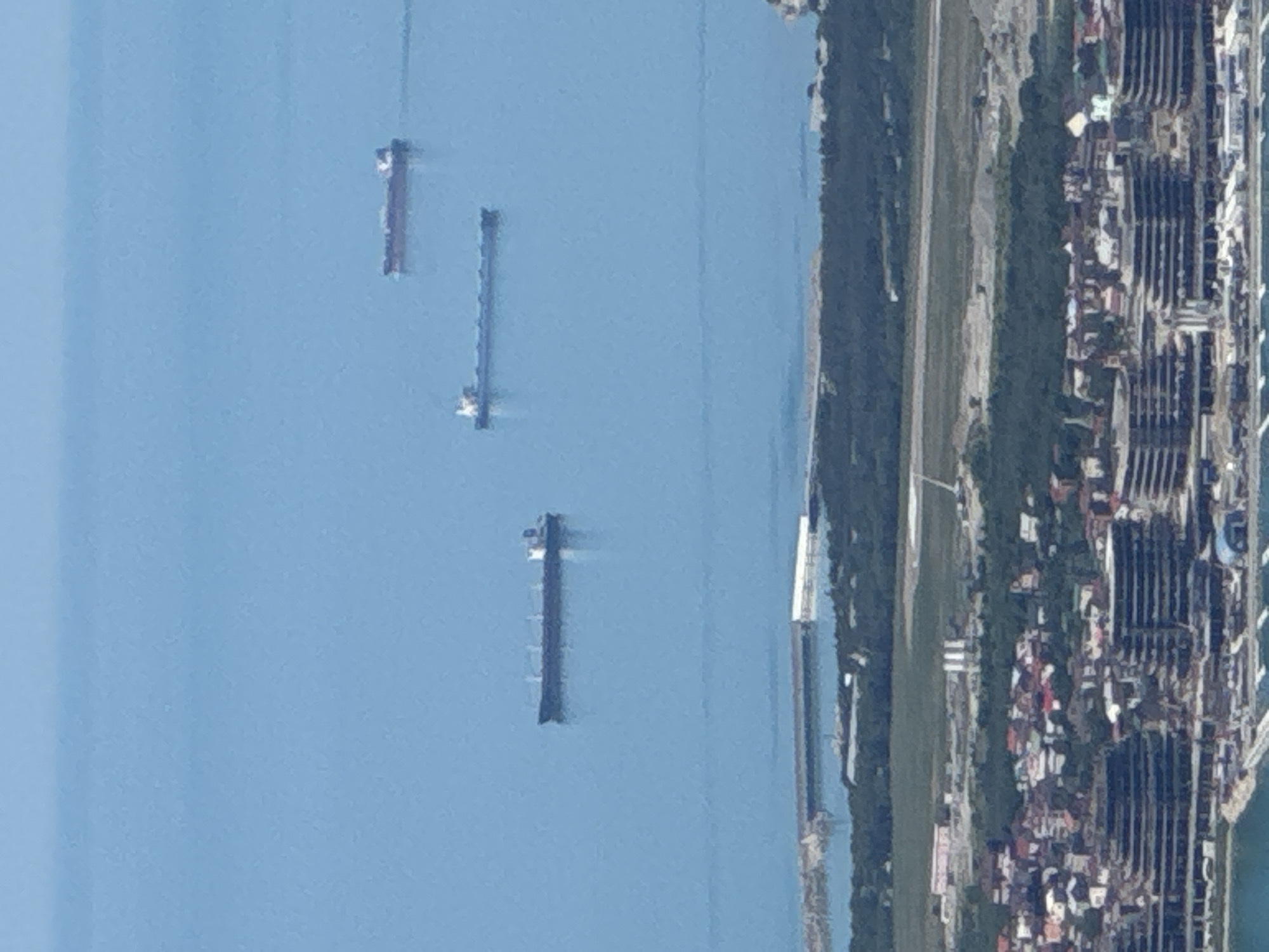 An aerial or high-angle photograph depicts a coastal landscape in Gelendzhik, Russia, under bright daytime conditions. The left side of the image features a wide expanse of calm, light blue water, likely the Black Sea. Three large, dark-hulled ships, possibly cargo vessels or tankers, are visible on the water, appearing as long, slender forms spaced out from each other and the shore.
To the right, the coastline unfolds with distinct features. A narrow band of green vegetation, possibly trees or parkland, runs parallel to the water's edge. Adjacent to this green strip is a lighter-colored, straight line, suggesting a road or a cleared path. Further inland, a densely built-up urban area is prominent, characterized by numerous, regularly arranged structures that appear to be large buildings, potentially residential complexes or hotels typical of a resort city. The overall scene suggests a developed coastal area with active maritime traffic. No people or visible text are discernible in the image.