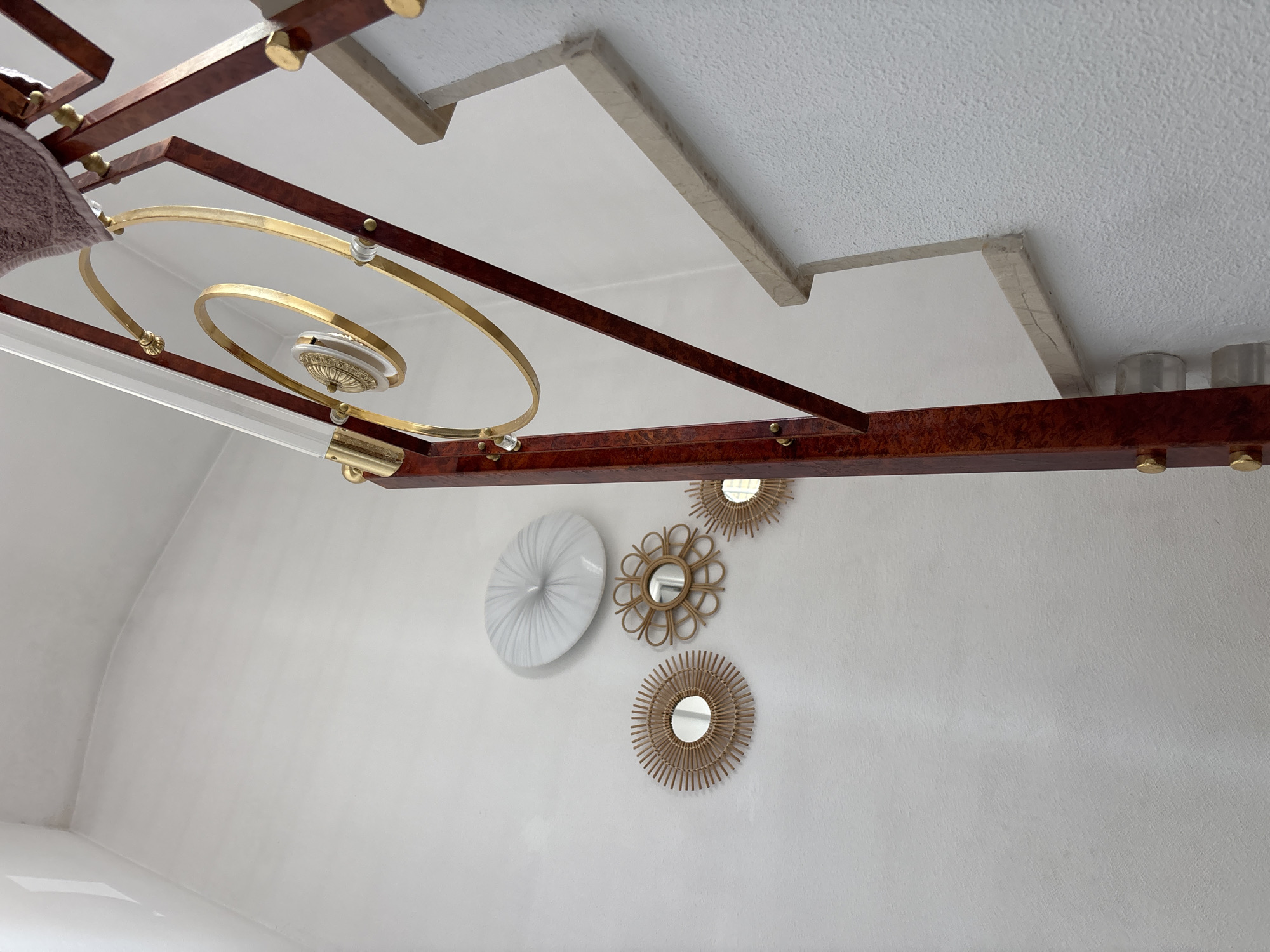 This media file captures an interior scene looking upwards towards a ceiling and a wall. The ceiling features a textured, white surface with several architectural details, including recessed areas with beige trim. Suspended from the ceiling is a decorative, multi-tiered lighting fixture. This fixture has a dark, polished wood frame with brass accents and includes gold circular elements and clear acrylic rods.
On the wall below, there are several decorative items. A large, round, white object with a pleated texture is mounted on the left. To its right, there are three round decorative mirrors with rattan frames. One mirror has a sunburst design, and the other two have a starburst or floral-like pattern.
The lighting suggests it is daytime, as natural light seems to be present, although the primary illumination comes from the fixture. There are no people visible in the frame. The overall impression is one of a well-decorated interior space, possibly a living room or dining area, with a modern and somewhat ornate aesthetic. The location context of Orihuela, Spain, does not provide specific visual cues within the image itself. No text is visible.