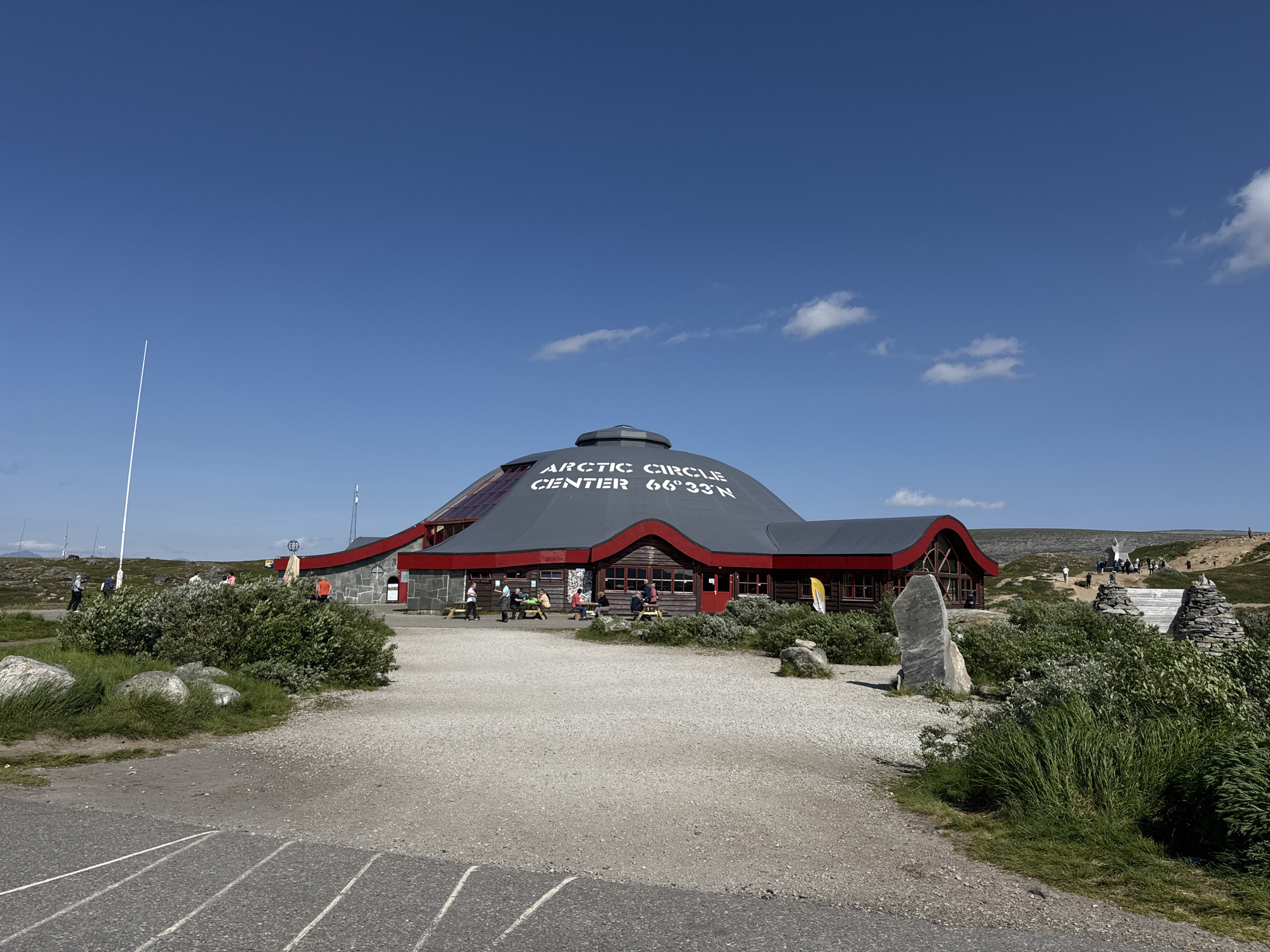 The image shows the Arctic Circle Center at 66°33' N, located in the vicinity of Storforshei, Norway. The building features a large, grey, domed roof with the text "ARCTIC CIRCLE CENTER 66°33' N" prominently displayed in white. The lower part of the building is red with wooden window frames and doors. Several people are visible around the building, some sitting at picnic tables outside. The surrounding environment is a barren, rocky landscape with low-lying vegetation, characteristic of a subarctic region. A gravel path leads to the building, and a tall white pole stands to the left. In the background, the terrain slopes upwards with more sparse vegetation and distant figures can be seen walking. The sky is clear blue with a few scattered white clouds.