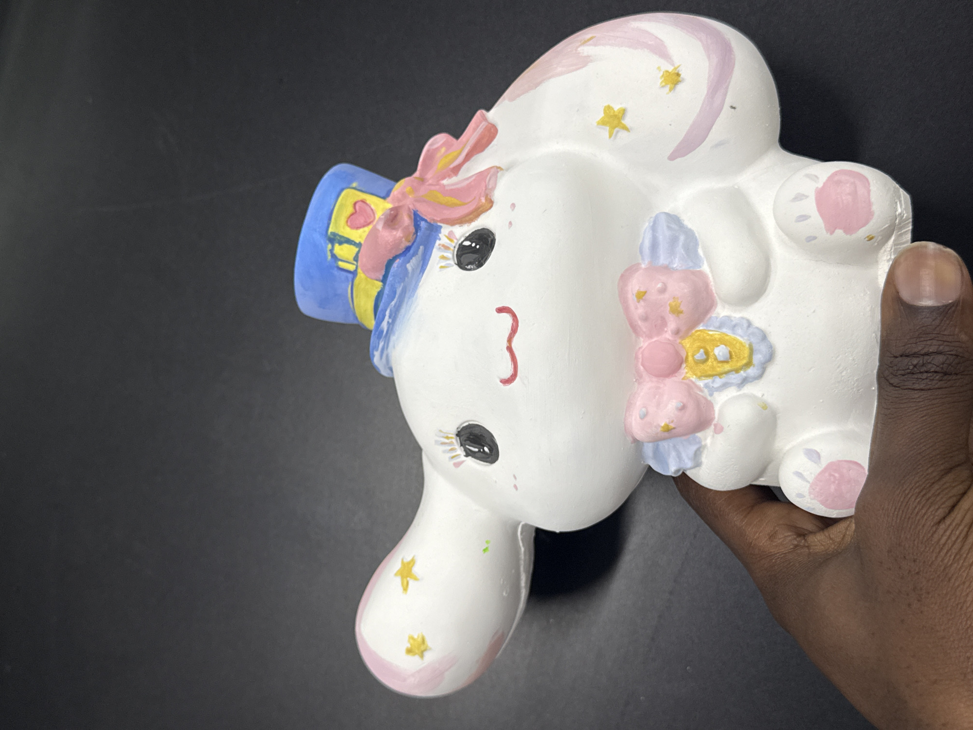 A person is holding a painted ceramic bunny figurine. The figurine is predominantly white, with pink, blue, and yellow accents. It features a hat, bows, stars, and a stylized "3" for a mouth. The background is a solid, dark color.