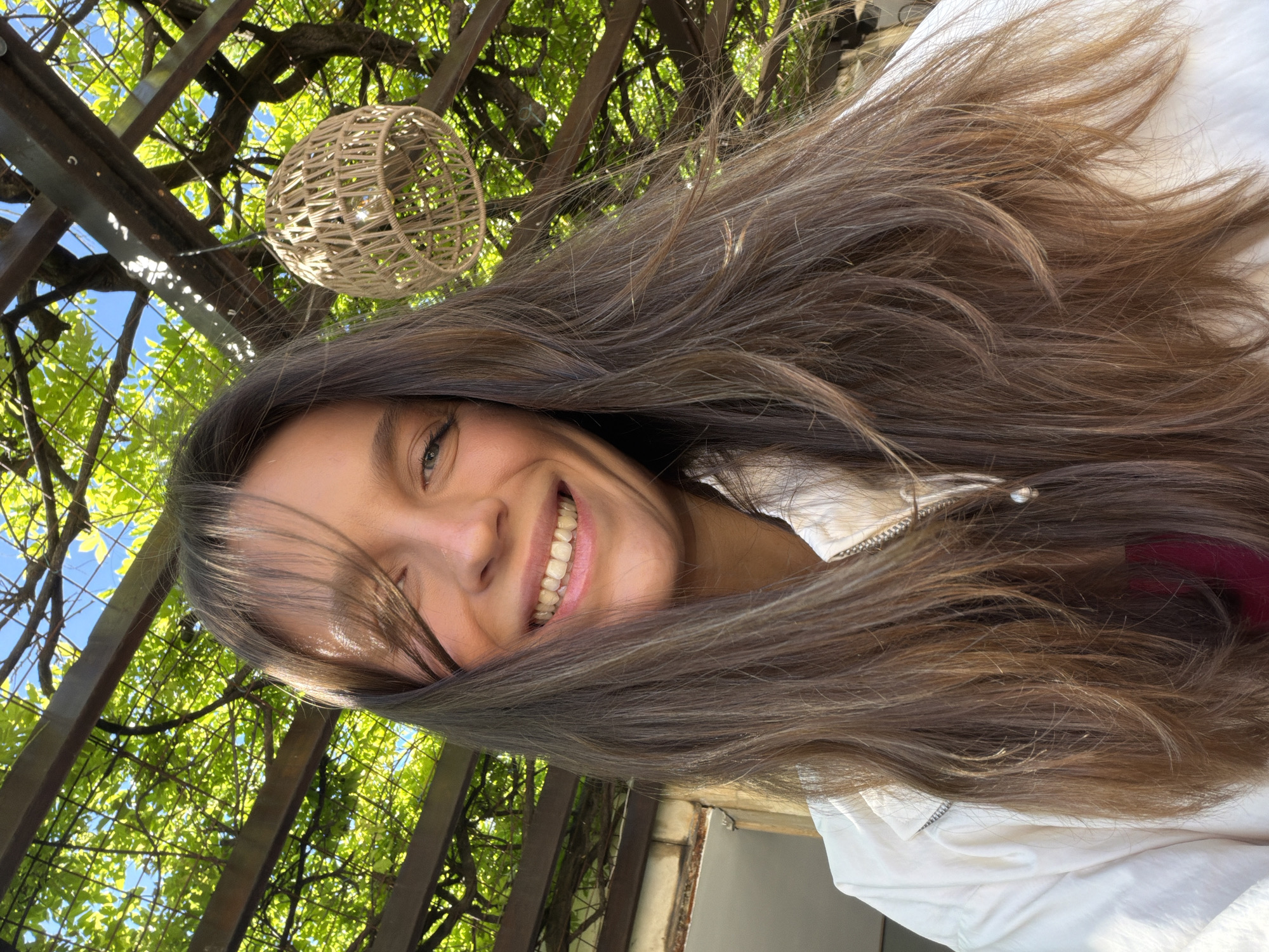 The image is a selfie taken from a low angle, showing a woman with long brown hair smiling. The background features a pergola-like structure with green foliage, hinting at a garden or outdoor space. A decorative woven ball is suspended from the pergola. The lighting is bright, suggesting a sunny day. Based on the context of Budva, Montenegro, the location could potentially be a garden or patio area of a restaurant or private residence.