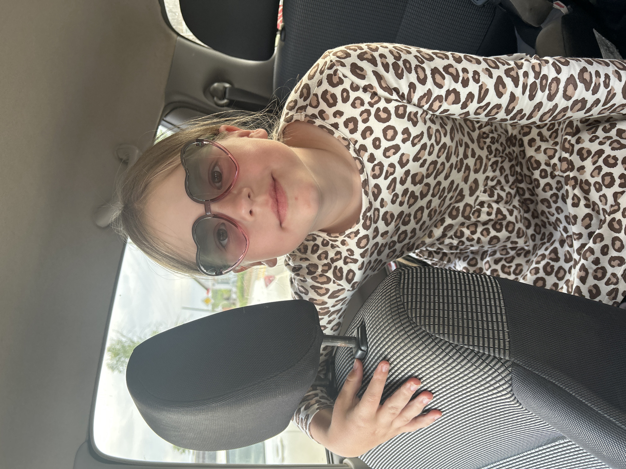 A young girl wearing heart-shaped sunglasses and a leopard-print top is in a car. She is positioned near the back of a front seat, her hand resting on the seat. The environment visible through the car window suggests an outdoor setting. The location, Фоки, Russia, cannot be confirmed from the visual information.