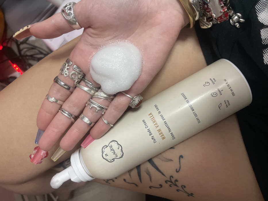 A person's hand, adorned with numerous silver rings and a bracelet, holds a dollop of fluffy white foam. A can of "Laline Warm Vanilla Fluffy Body Cream" rests against the hand. The can highlights the cream's silky soft moisture boost and ingredients like Aloe Vera extract and Vitamin E. A tattoo is visible on the person's leg. The can’s volume is marked as 200 ml or 6.76 fl. oz.