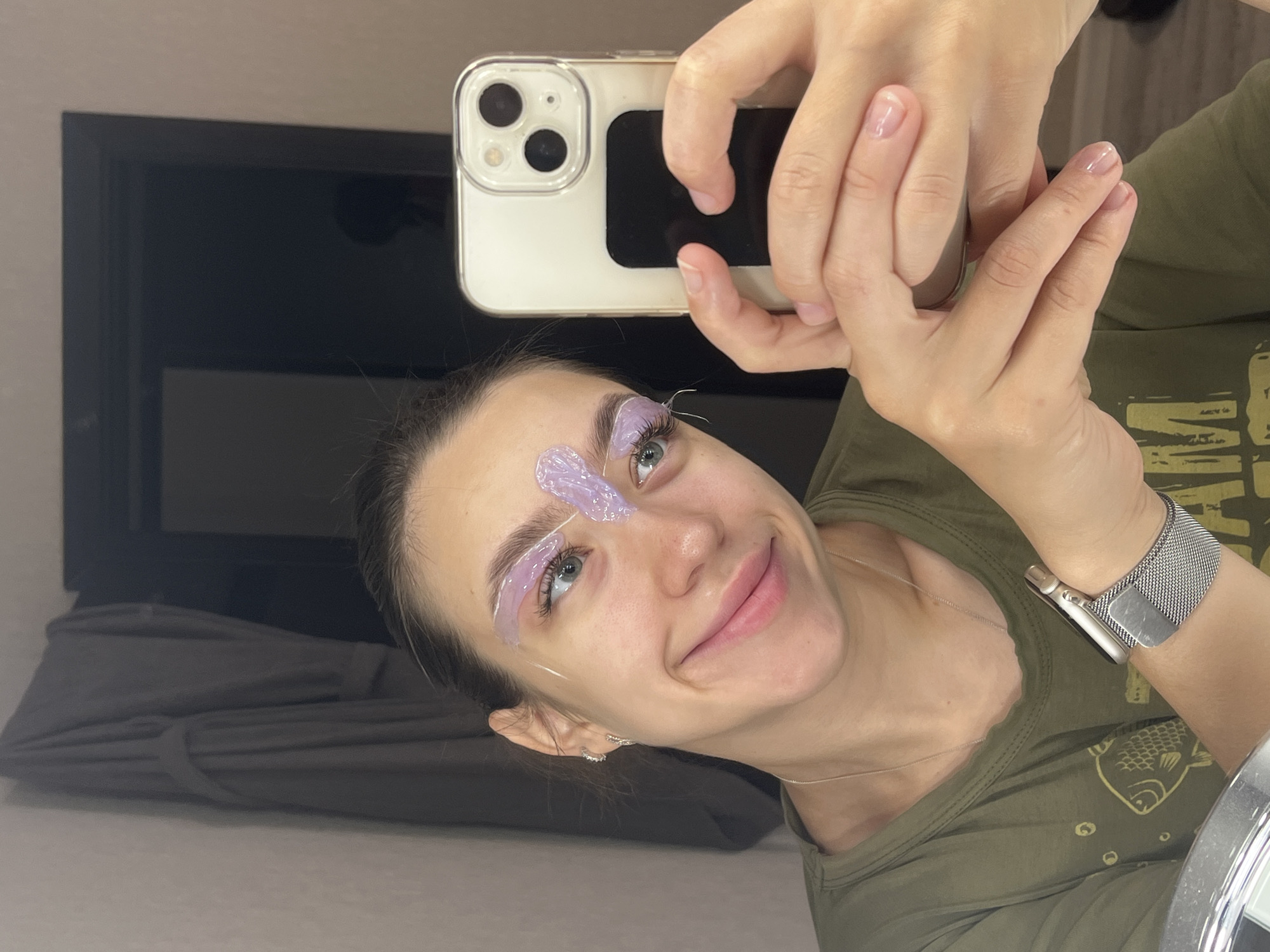 A young woman with dark hair pulled back is looking into a smartphone held in her hands, taking a selfie. She is smiling and has a translucent purple substance applied to her eyebrows, possibly for tinting or shaping. She is wearing a green t-shirt with a yellow fish graphic. A silver smartwatch is on her left wrist. The background appears to be indoors, with a gray bathrobe hanging on the left and a dark doorway or cabinet on the right. The scene appears to be a personal moment captured in a home environment. The location is Chaikovsky, Russia.