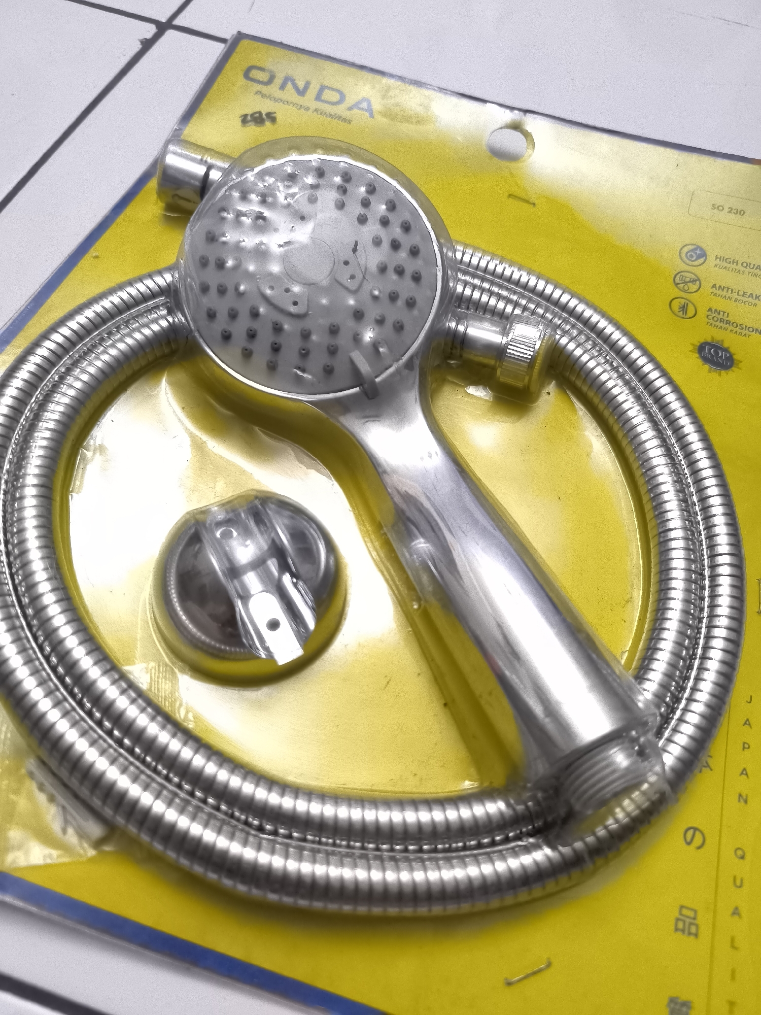 An overhead close-up shot shows a new silver handheld showerhead with a flexible hose, packaged on a bright yellow card that is partially on a white tiled surface. The showerhead, made of metal, has a circular face with multiple small holes. A coiled silver hose connects to the base of the showerhead. A small, clear plastic holder for the showerhead is also visible. The yellow packaging features a brand logo that reads "ONDA" in large blue letters, along with smaller text and icons indicating product features such as "HIGH QUALITY" and "ANTI-CORROSION". The background shows a white tiled floor with dark gray grout lines.