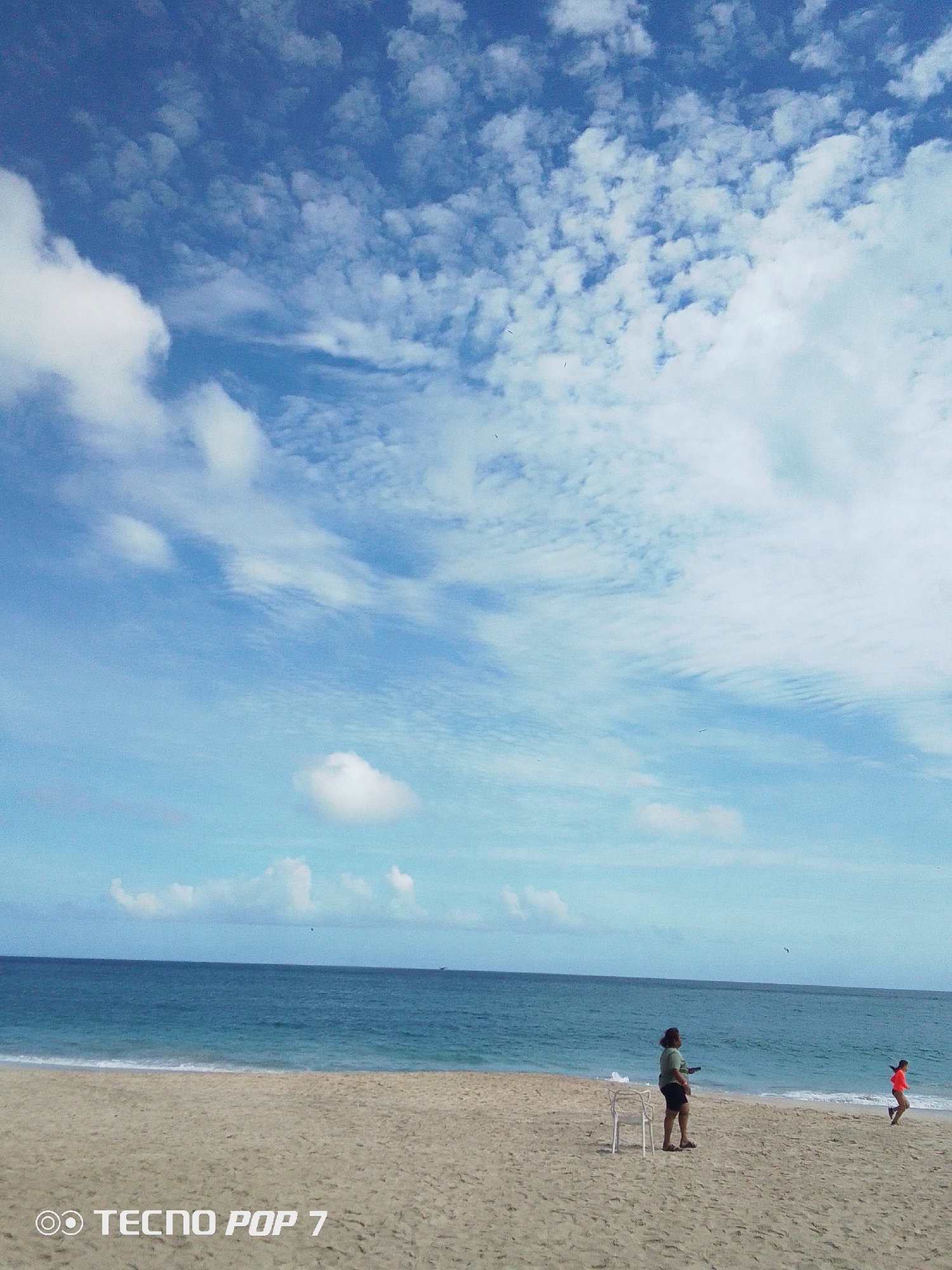 The image depicts a beach scene in Pedro González, Venezuela. The sky is mostly blue with scattered cumulus clouds. The sea is a darker shade of blue. The beach is sandy, and a few small waves are visible along the shoreline.
A woman stands on the beach near a white plastic chair. She is wearing a light-colored shirt and dark shorts. A child in an orange shirt runs along the shoreline.
