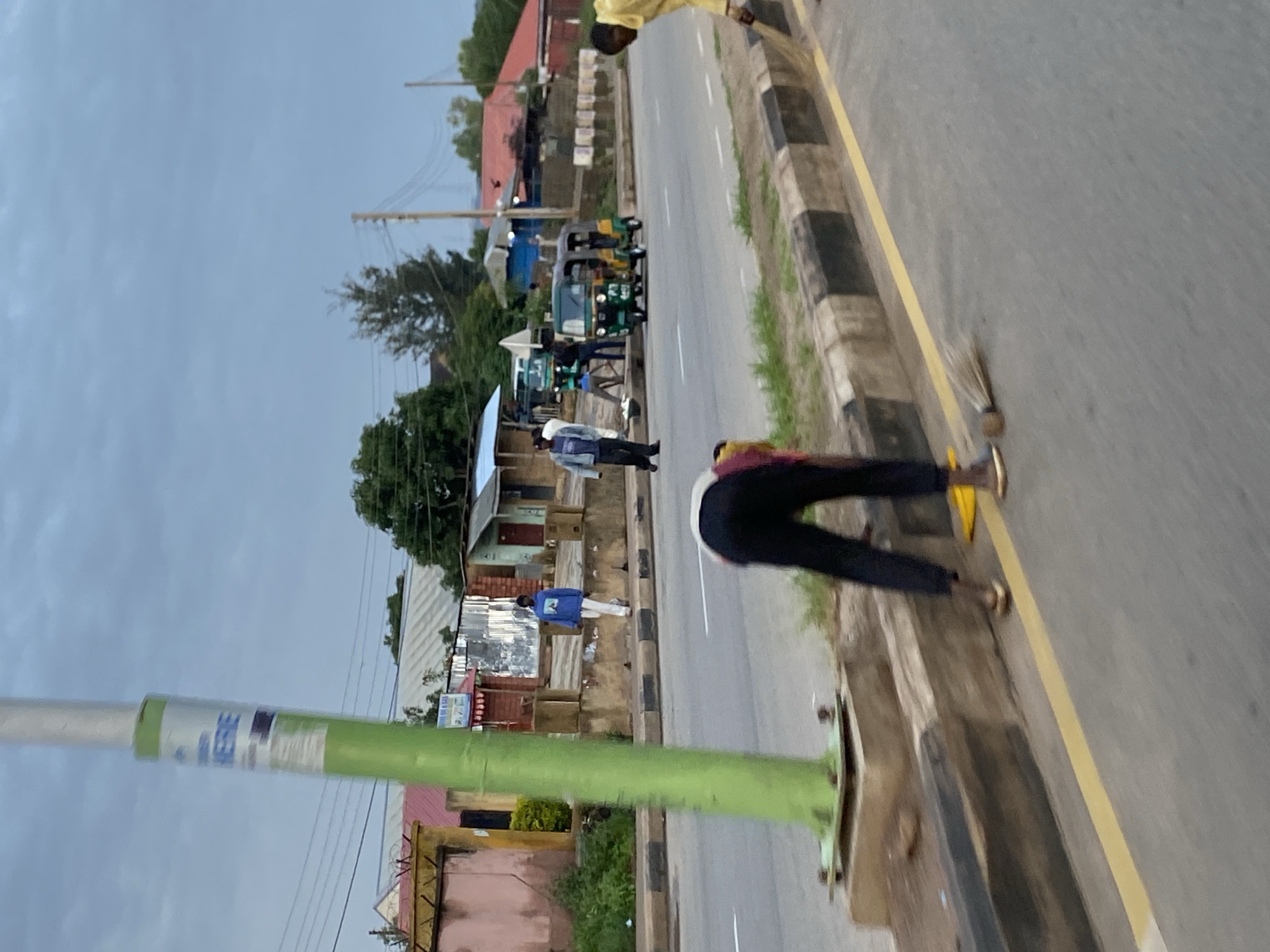 The image depicts a roadside scene in Jos, Nigeria, under an overcast sky. A person is sweeping the street, with a broom and a yellow dustpan visible. The street has a yellow line painted along the curb, which is lined with black and white checkered patterns. In the background, several people are standing near a road with buildings and some vegetation. One building has a sign that says "HOTEL". A green and yellow tuk-tuk, a common mode of transportation in Nigeria, is parked on the side of the road, with people near it. A green lamppost stands prominently in the foreground. The overall scene suggests a typical day in a bustling urban area, with activity focused around the street and nearby businesses.