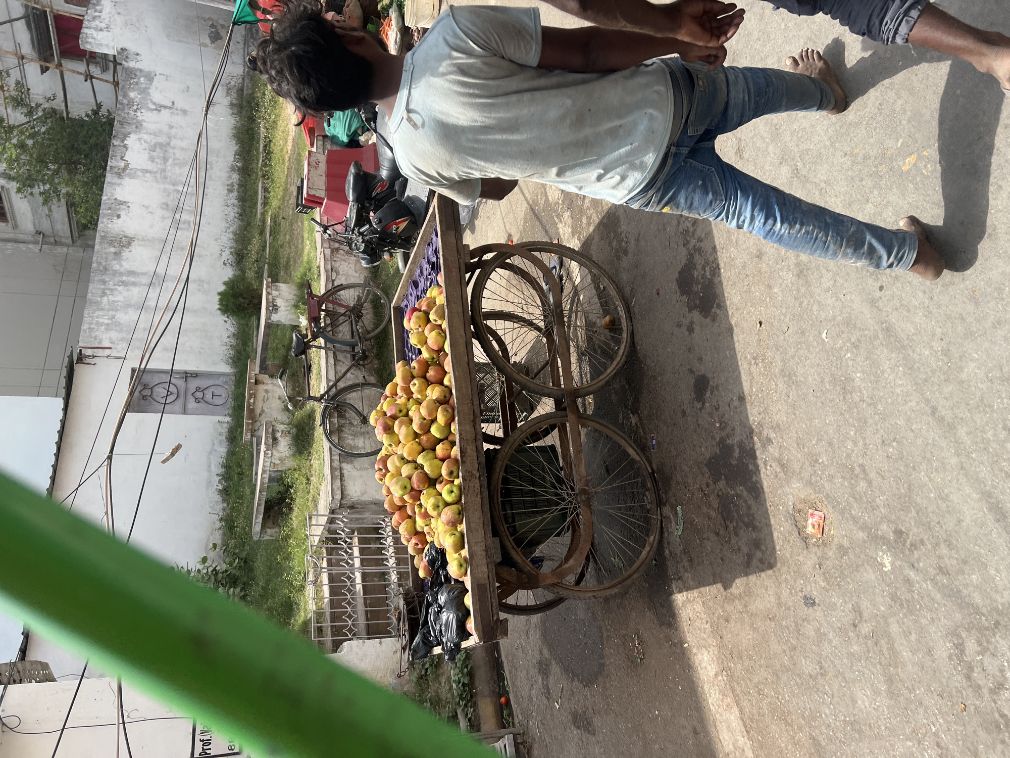 The image shows a street scene in Varanasi, India. An Indian man, wearing a light-colored t-shirt and blue jeans, is standing near a cart laden with apples. He is barefoot. Another person, only partially visible, stands next to him. The cart has four wheels and is heavily filled with apples. There are bicycles parked in the background, along with some greenery and buildings. The ground appears to be paved, and the scene is bathed in bright sunlight.