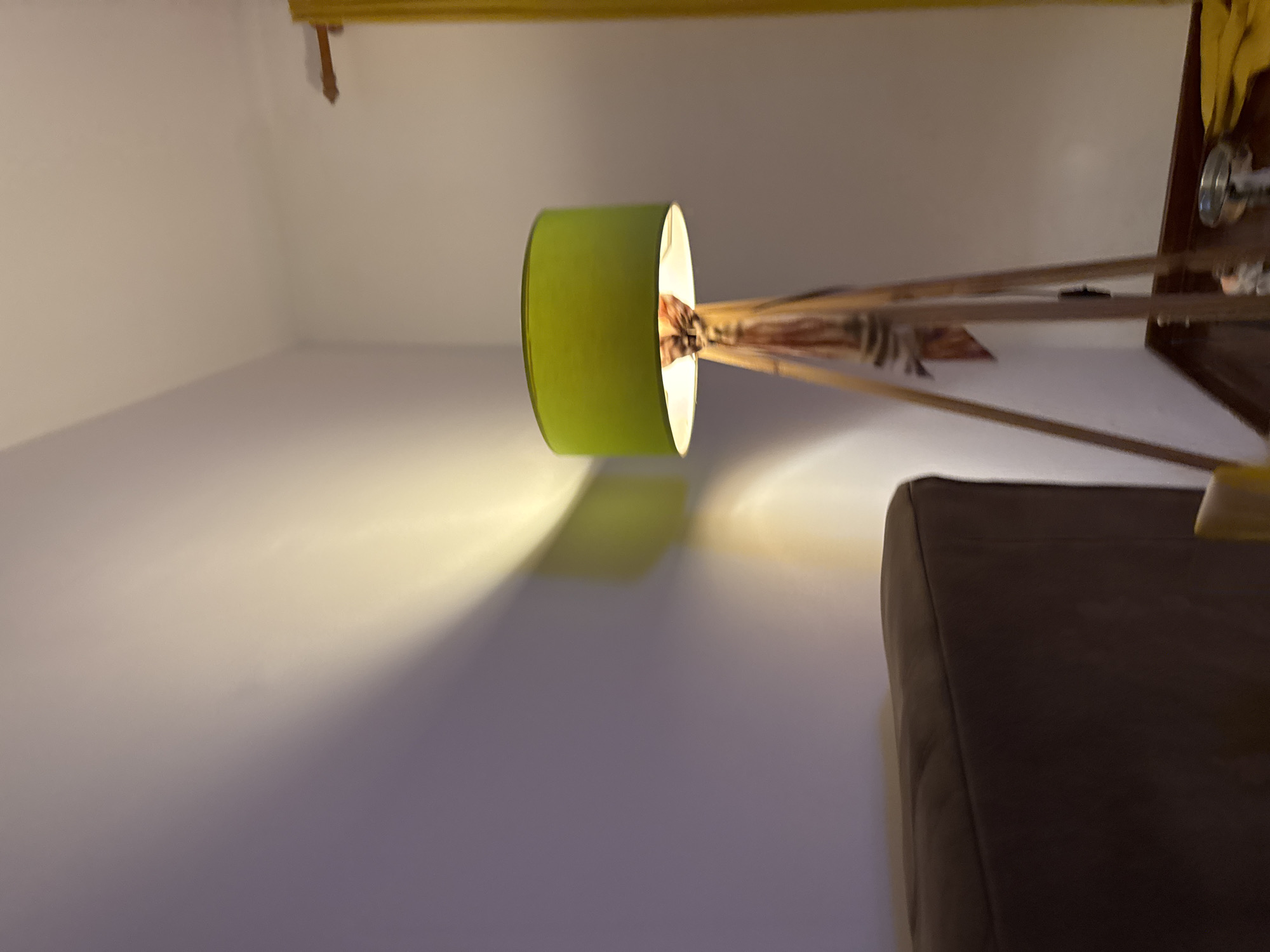 The image depicts a room interior featuring a floor lamp with a green lampshade casting a beam of light onto a white wall. The lamp appears to have a wooden tripod base. A dark brown sofa is positioned on the right side of the image. Above, a yellow curtain rod is visible near the ceiling. The location is not discernible from the visual information provided. The photograph captures a static scene with no visible human presence or active interactions.