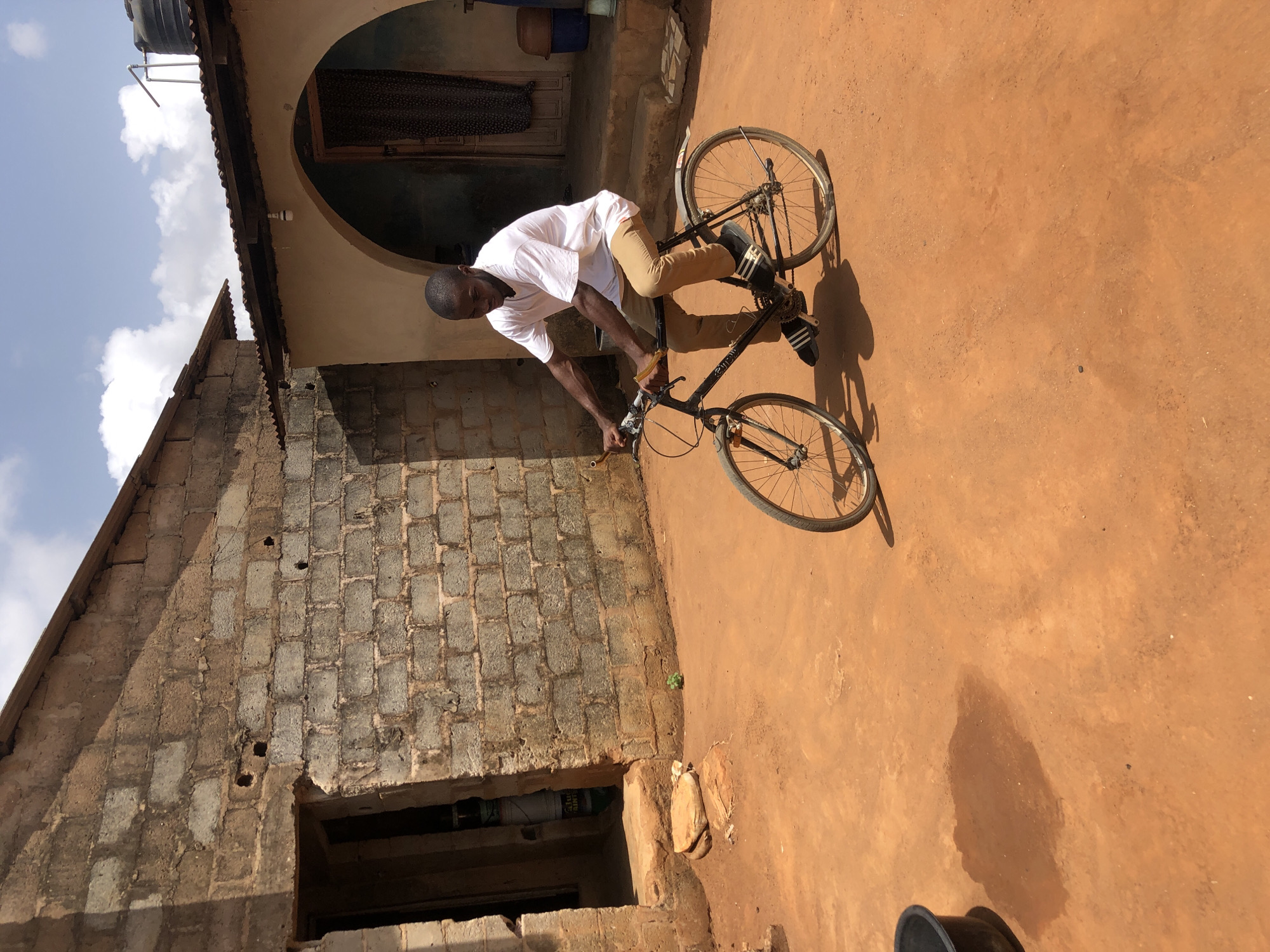 A man wearing a white t-shirt and light brown pants is riding a bicycle in an outdoor area. The ground is a reddish-brown color, and the man is looking down at the handlebars of the bicycle. A building made of gray blocks is to the left of the man. The building has a window near the bottom and a doorway above. To the right of the man, there is a doorway with an arch over it. The location is Unknown, Unknown.