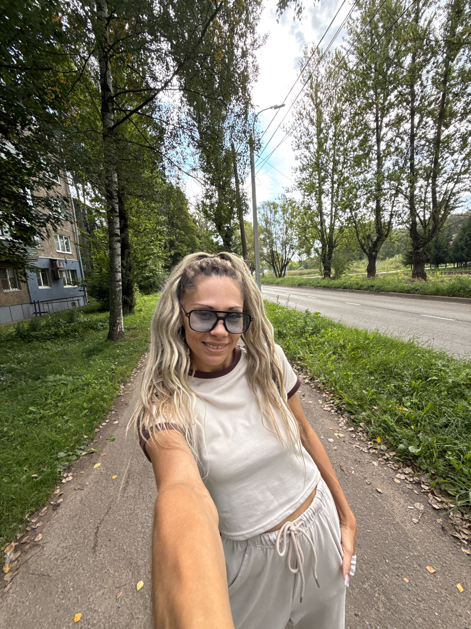 A woman with long, blonde dreadlocks and sunglasses is taking a selfie outdoors. She is wearing a beige t-shirt and matching sweatpants with a drawstring. She is standing on a paved walkway next to a grassy area. In the background, there is a multi-story apartment building on the left and a road with trees lining it on the right. The sky is overcast. The location is identified as Rusanovo, Russia.