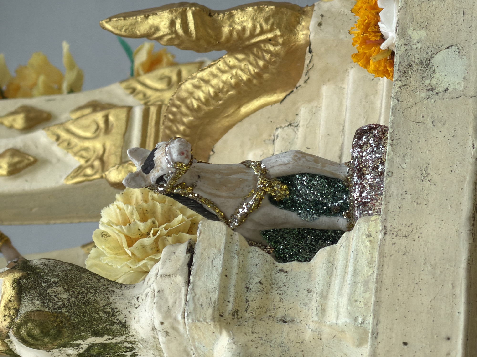 The image shows a close-up of a small, white statue of a horse with black markings on its face. The horse is adorned with a gold glittery necklace and halter. It stands on a base covered in green glitter and what appears to be a mound of red and silver glitter. Behind the horse is a larger, ornate structure with gold detailing, possibly a shrine or altar. There are yellow flowers in the background and orange flowers in the foreground. The overall scene suggests a religious or decorative display. The location is Unknown, Unknown.