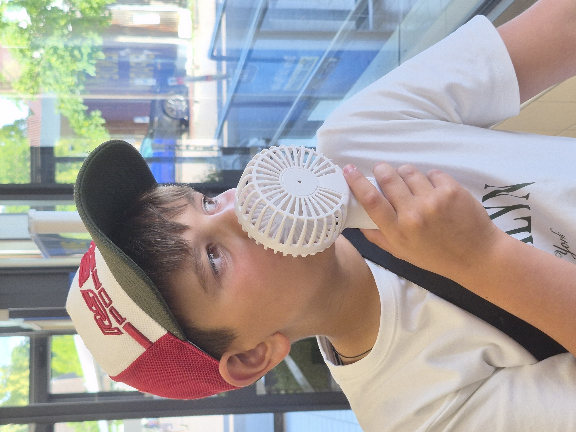 A young boy wearing a red, white, and green baseball cap and a white t-shirt is holding a small, white portable fan up to his face. He is looking to his right, away from the camera. The fan is circular with visible spokes. The boy has brown hair and brown eyes. He is wearing a thin dark necklace. In the background, there is a large window with a reflection of trees and buildings, and a street scene with cars and other people visible outside. The setting appears to be indoors, possibly a shop or restaurant, given the window and reflections.