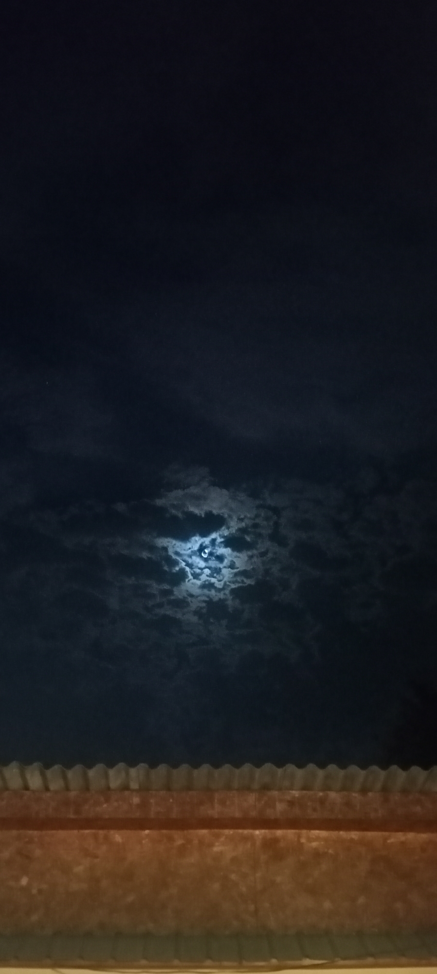 An overcast night sky is visible above a textured surface, likely a corrugated metal roof. The moon is partially obscured by clouds, casting a faint glow. A dark, rough-textured wall is in the foreground, with the suggestion of a green structure below. The scene appears to be from a residential area in Sokuluk, Kyrgyzstan.