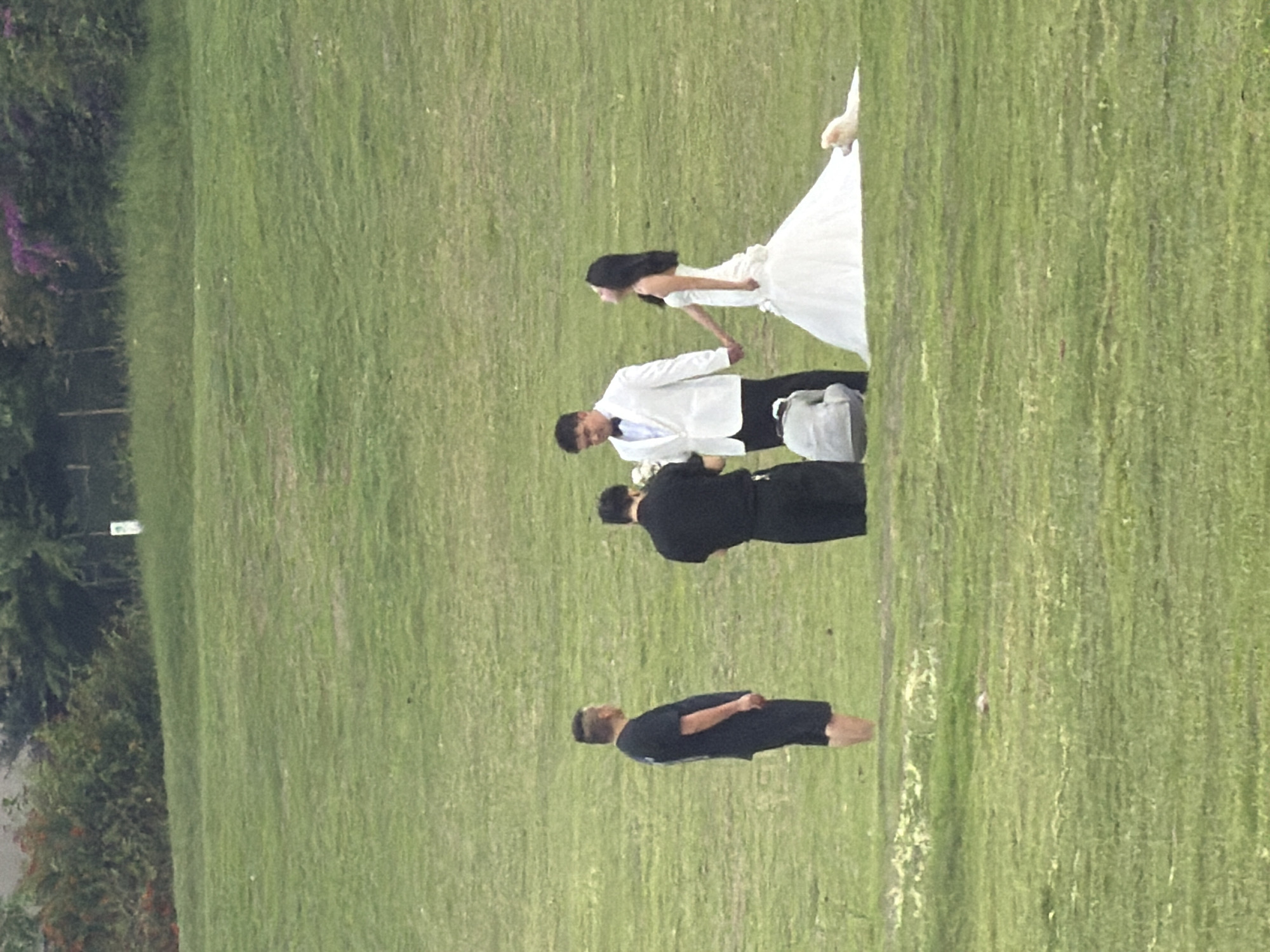 In Jiangjin District, Chongqing, China, on a grassy hillside, a wedding photoshoot is taking place. A man in a white suit and a woman in a white wedding dress are holding hands. A person in a black outfit is standing to the left of the groom, and another person in black is kneeling in front of them, possibly as a photographer or assistant. A light-colored dog is lying on the grass near the bride. The background features a line of trees and foliage, with a small white marker visible in the distance. The overall environment is natural and open, with the focus on the wedding party.