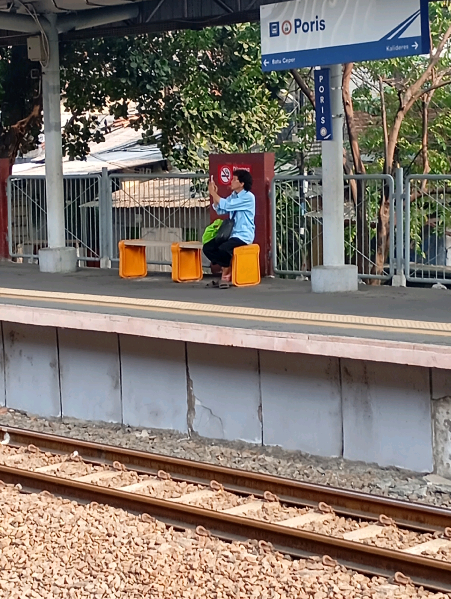 The image depicts a platform at the Poris train station in Tangerang, Indonesia. A person, wearing a light blue shirt and dark pants, is seated on an orange bench and appears to be taking a photograph with their phone. The platform is bordered by train tracks on the foreground and a concrete wall behind the bench. A sign for "Poris" is visible overhead, with directional arrows indicating routes to "Batu Ceper" and "Kalideres." Trees and foliage are present in the background, suggesting an outdoor setting. The overall scene is a snapshot of daily life at a public transportation hub.