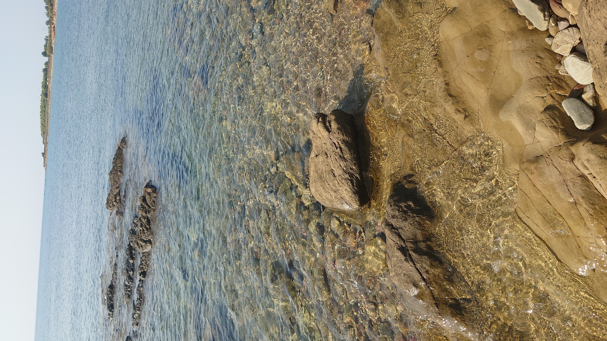 This image depicts a rocky coastline at a body of water, likely the sea, with a clear, shallow area revealing pebbles and submerged rocks. In the foreground, several large, tan-colored rocks are visible, some partially submerged in the water. The water's surface is disturbed by gentle ripples, creating patterns of light and shadow on the seabed. In the middle ground, darker, more rugged rocks jut out of the water, appearing as small islands or submerged formations. The background shows a vast expanse of calm, blue water under a clear sky. In the far distance, a thin strip of land with vegetation and possibly structures is visible along the horizon. The scene appears to be a natural coastal environment, likely near a beach or shoreline, consistent with the coastal geography of Villaputzu, Italy. There are no people or animals present in the image.