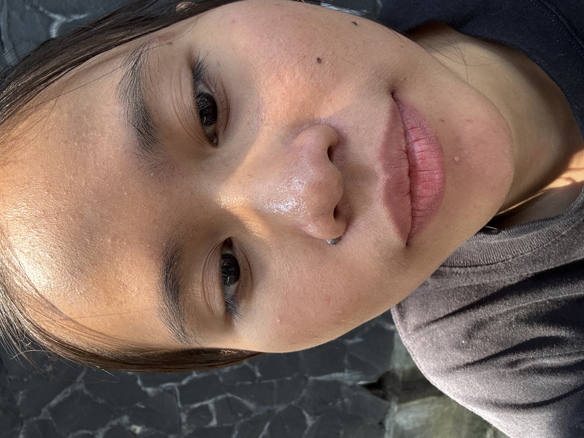 A close-up, low-angle selfie captures a young woman's face outdoors against a dark, textured stone wall, likely located in South Jakarta, Indonesia. Sunlight highlights her forehead and the bridge of her nose, creating a subtle sheen on her skin. She has dark hair pulled back, revealing her forehead and the upper part of her face. Her eyes are dark, and her eyebrows are naturally shaped and filled. A small stud pierces her left nostril. Her lips are slightly parted, revealing a healthy pink hue. Her skin shows a few blemishes and appears to have some texture. She is wearing a dark grey t-shirt. The overall lighting suggests it is daytime, possibly mid-morning or late afternoon, given the angle of the sun.