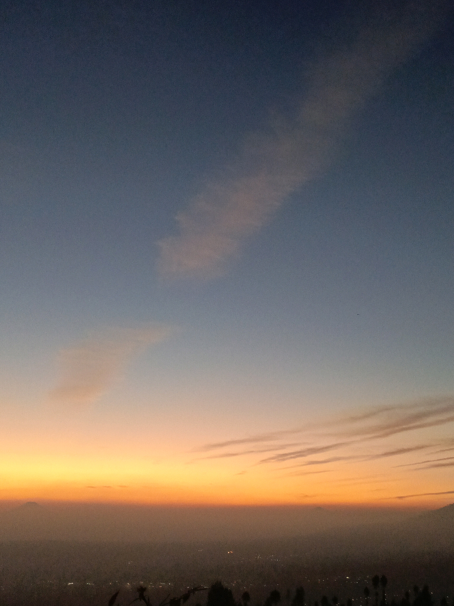 A wide shot captures a hazy, distant view of Butuh Kidul, Indonesia, at dusk. The sky transitions from a deep blue at the top to a vibrant orange and yellow near the horizon, illuminated by the setting sun. Wispy clouds streak across the sky, catching the fading light. Below, the city is shrouded in a thick, brownish haze, with faint, scattered lights indicating its presence. The foreground is dominated by dark silhouettes of trees and foliage, suggesting the photo was taken from an elevated vantage point.