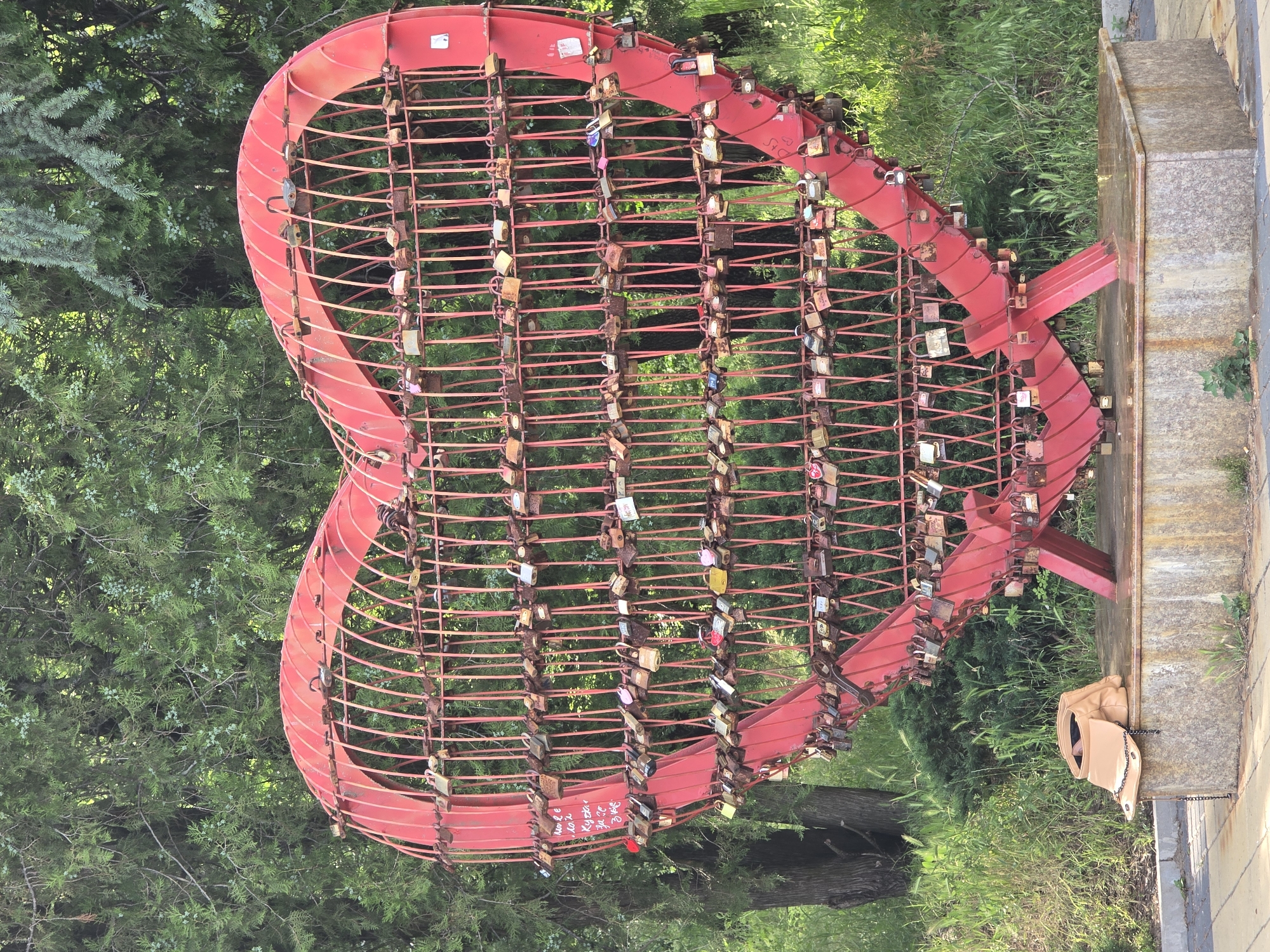 A medium, slightly-wide, eye-level shot shows a large, red, heart-shaped metal cage covered in locks in a park in Unknown, Unknown.
The heart is made of thick metal bars shaped to form a heart. Inside the heart shape are thinner, horizontal metal bars that create a grid. Locks of various sizes and colors are attached to the bars, densely populating the structure.
The heart is mounted on a platform made of stone or concrete. A small tan handbag hangs off the side of the platform, secured by a metal chain.
Behind the heart is dense green foliage, including bushes and trees. A single tree trunk is visible between the bottom point of the heart and the platform. The light is bright and diffused, suggesting an overcast day.