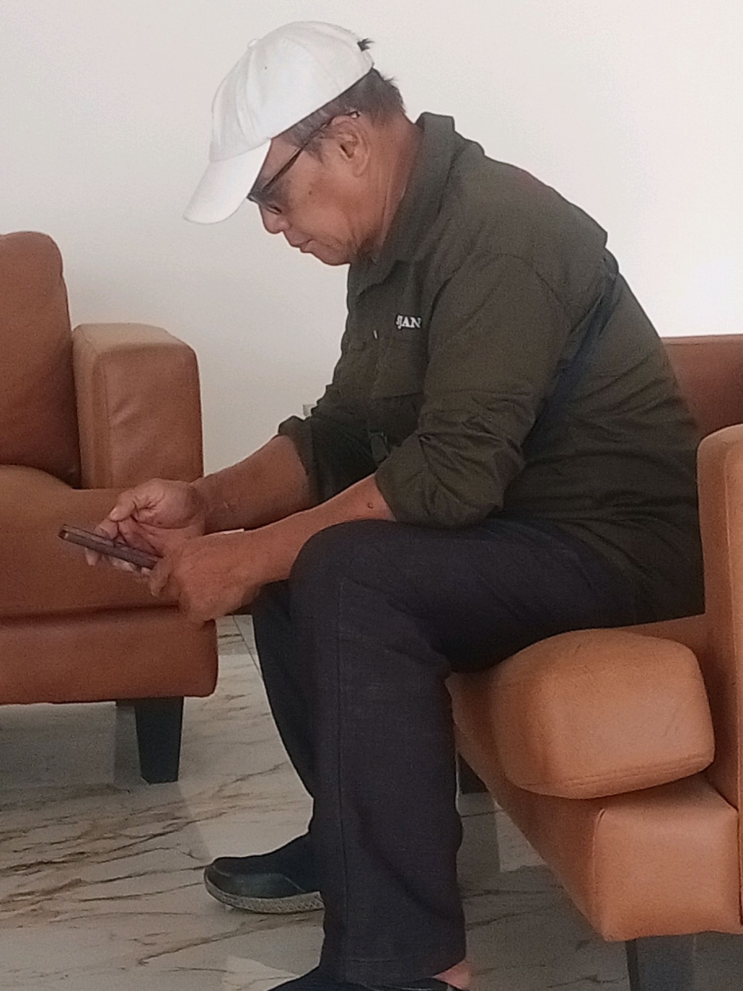 A middle-aged man wearing a white baseball cap and glasses sits on a brown leather sofa, looking at his phone. He is wearing a dark olive green collared shirt with "BAN" visible in white lettering on the left breast and dark navy pants. His feet are clad in black slip-on shoes. The sofa is in a room with a white wall and a marble-patterned floor. The scene appears to be a waiting area or a casual living space, possibly within a commercial building or a home in Samarinda, Indonesia. The lighting suggests an indoor environment, likely during the daytime.