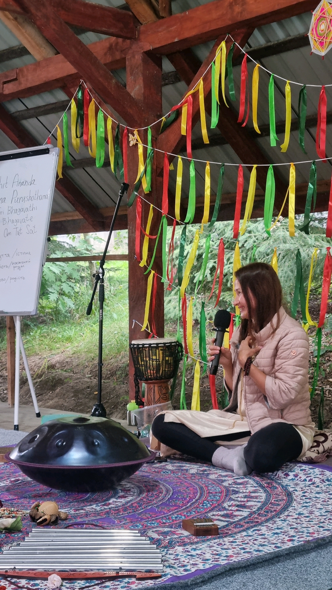 A woman is sitting cross-legged on a circular rug in what appears to be an outdoor, rustic setting, possibly a covered wooden pavilion. She is holding a microphone and speaking, with a warm smile. She is wearing a light pink puffer jacket over a cream-colored garment and black leggings with white socks. In front of her, on the rug, is a large, dark, metallic handpan drum. To the left of the handpan, a microphone stand is visible, and further left, a whiteboard with writing on it is propped up. To the right of the woman, a djembe drum sits on a decorative base. The background shows trees and foliage, suggesting a natural environment. Red, yellow, and green ribbons are strung across the wooden structure above. The location is specified as Izky, Ukraine, and the overall atmosphere suggests a workshop, performance, or spiritual gathering.