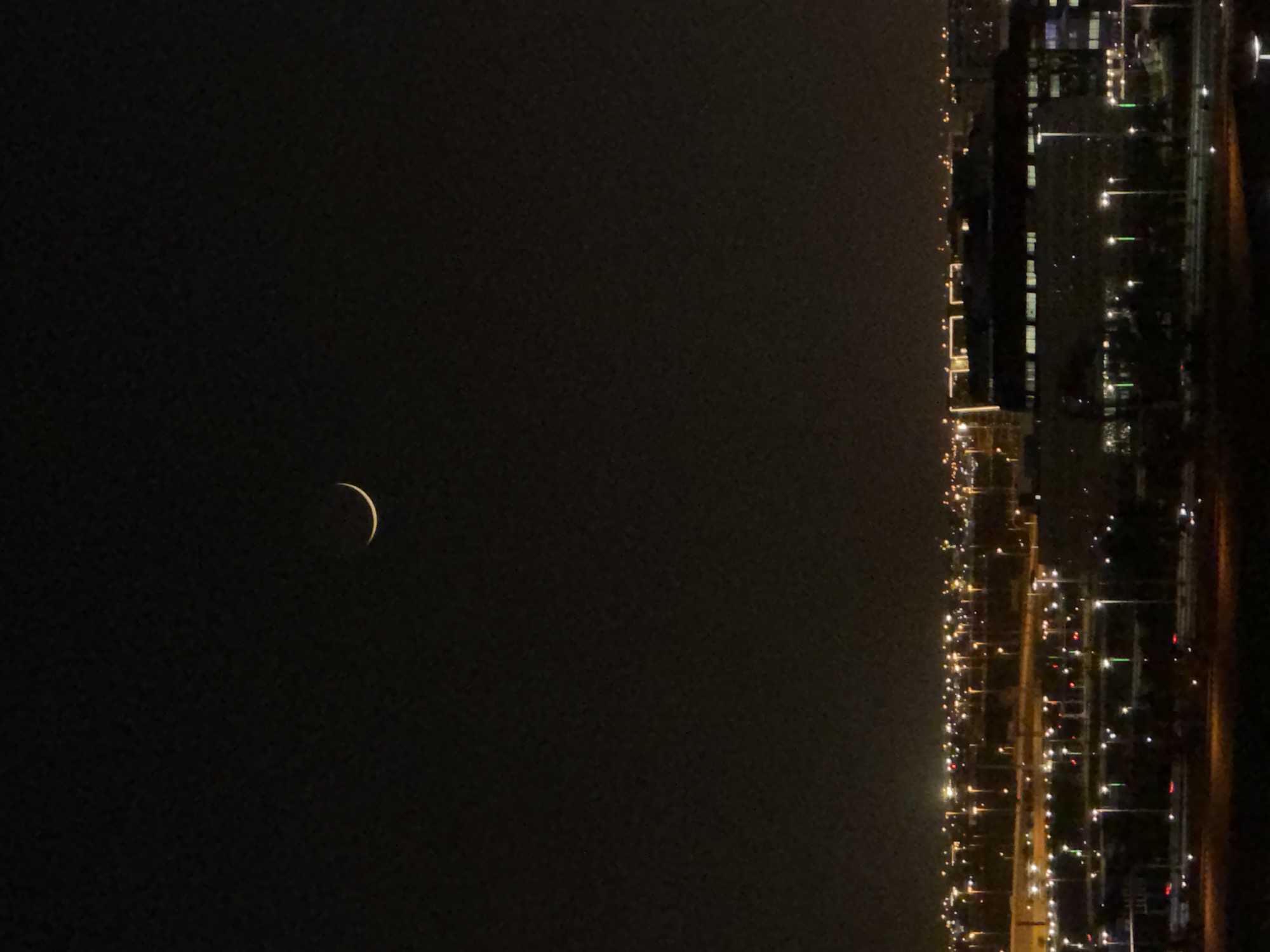 A crescent moon is visible in the night sky above a city. The city is illuminated by streetlights and building lights, creating a glow on the horizon. The foreground is dark, with the lights of the city extending into the distance.