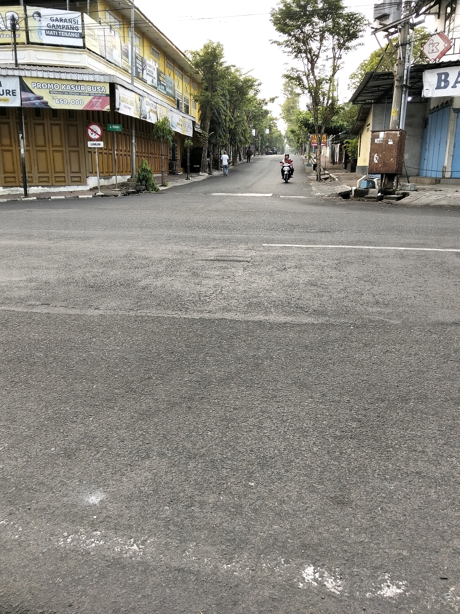 The image depicts a street scene in Purworejo, Indonesia, possibly in the early morning or late afternoon given the lighting. The street is paved and appears to be a commercial area with buildings lining the left side. These buildings are yellow and house various shops, indicated by signs for "GARANSI GAMPANG HATI TENANG," "PROMO KASUR BUSA," and "FURNITURE KANTOR." The storefronts on the left are mostly shuttered with wooden doors. A traffic sign with a red circle and a horizontal bar, likely indicating a prohibition, is visible near the sidewalk.
On the right side of the street, there are also buildings and some utility poles. One building has a blue facade and a sign with the letter "BA." A rusted metal utility box is mounted on a pole on the right side.
There are a few individuals present. A person in a white shirt is walking away from the camera on the left side of the street, and another person is riding a motorcycle on the right side of the street, moving away from the camera. The environment appears to be a typical urban street with trees lining the sidewalks. The overall impression is a quiet commercial street with limited activity.