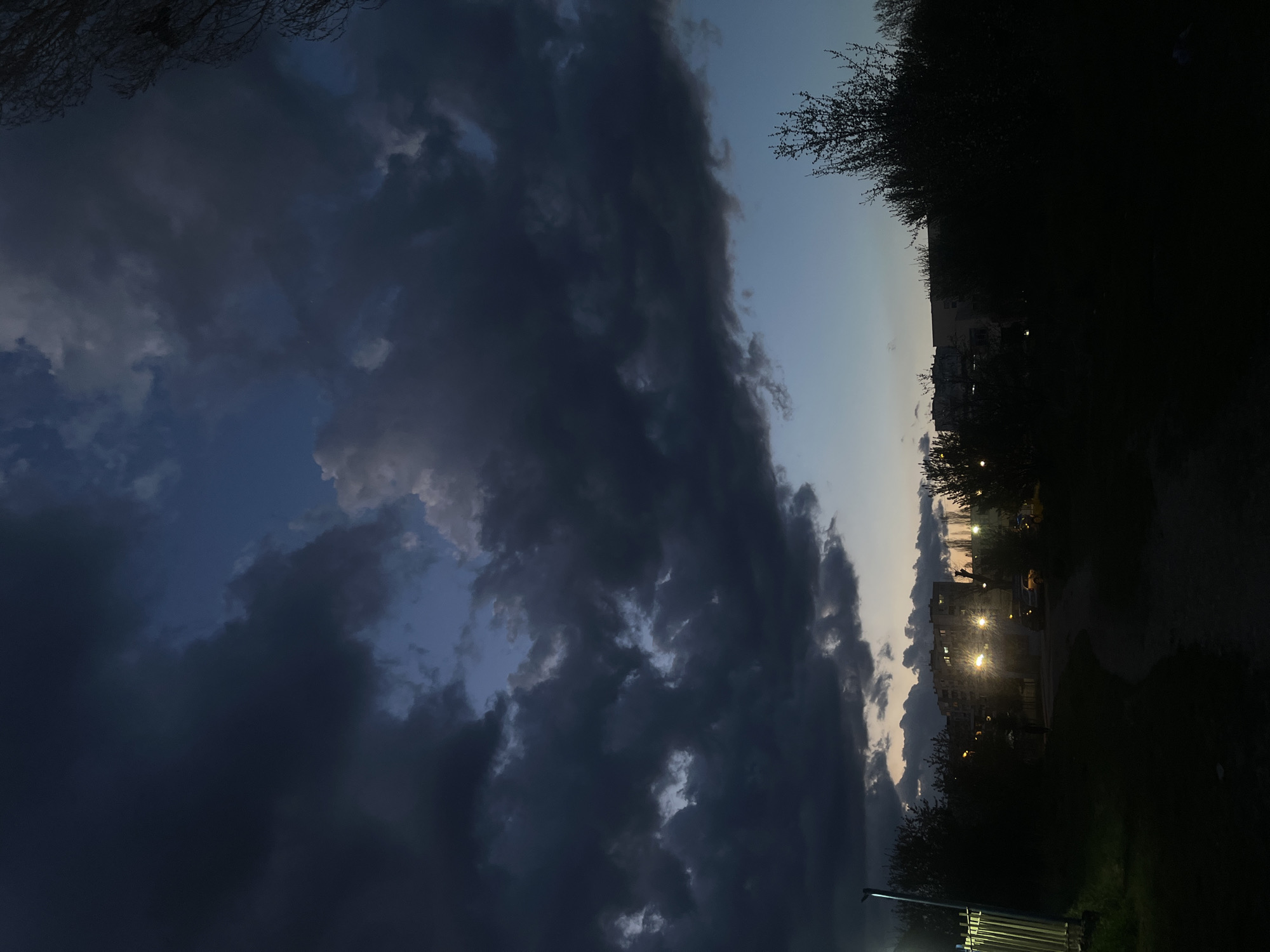 The image depicts a nighttime urban scene in Sofia, Bulgaria. Dark, heavy clouds dominate the left portion of the sky, contrasting with a lighter, almost twilight-like area towards the center. On the right side of the image, a dark, silhouetted area suggests trees and possibly a park. Further in the distance, illuminated buildings with lights are visible, indicating an urban setting. The foreground includes blurry outlines of foliage and structures, suggesting a low vantage point. Overall, the image captures a dramatic sky over a partially illuminated urban landscape at night in Sofia.