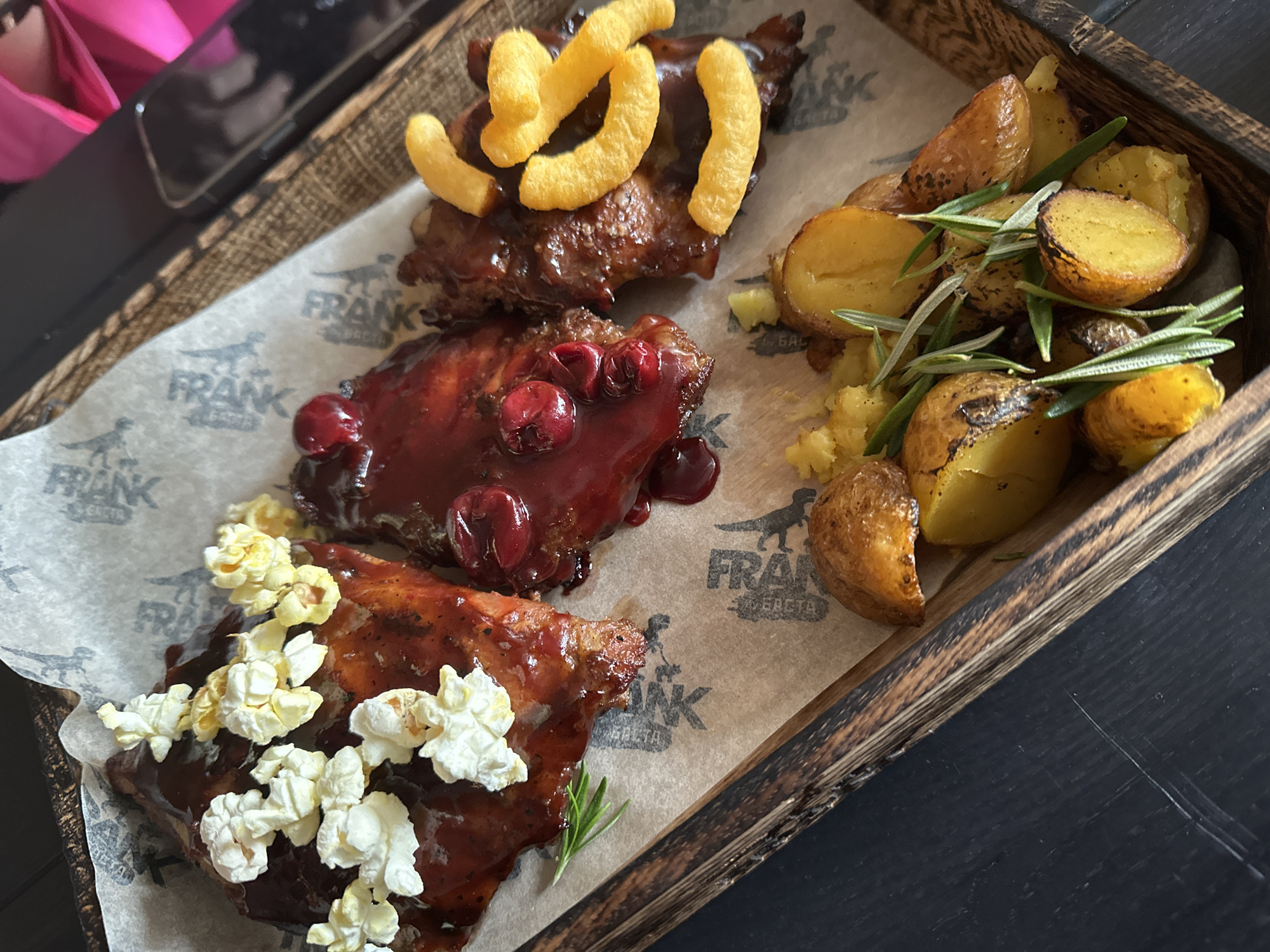 An overhead shot depicts a wooden serving tray containing a meal, resting on a dark, scratched surface. The tray is lined with light brown parchment paper, branded with a repeating dark grey logo featuring the word "FRANK" and an animal graphic, identifying the establishment as Frank Bar & Ribs in Saratov, Russia.
The meal consists of three distinct portions of sauced meat, likely ribs, and a side of roasted potatoes.
1. The leftmost meat portion is coated in a dark red-brown barbecue sauce and garnished with white and yellow popcorn. A sprig of fresh rosemary is positioned beneath it.
2. The central meat portion is covered in a glossy, dark red sauce and topped with several whole red cranberries or similar berries.
3. The rightmost meat portion is coated in a dark brown sauce and garnished with multiple bright orange, puffed, curved snack pieces, resembling cheese puffs.
Below these meat portions, a cluster of small, roasted potatoes, some halved to reveal yellow flesh, are arranged. They exhibit a golden-brown exterior and are interspersed with several sprigs of fresh rosemary.
In the upper right background, a black smartphone with a visible camera lens is partially in frame, adjacent to a brightly lit, out-of-focus pink object. No people are visible in the image.