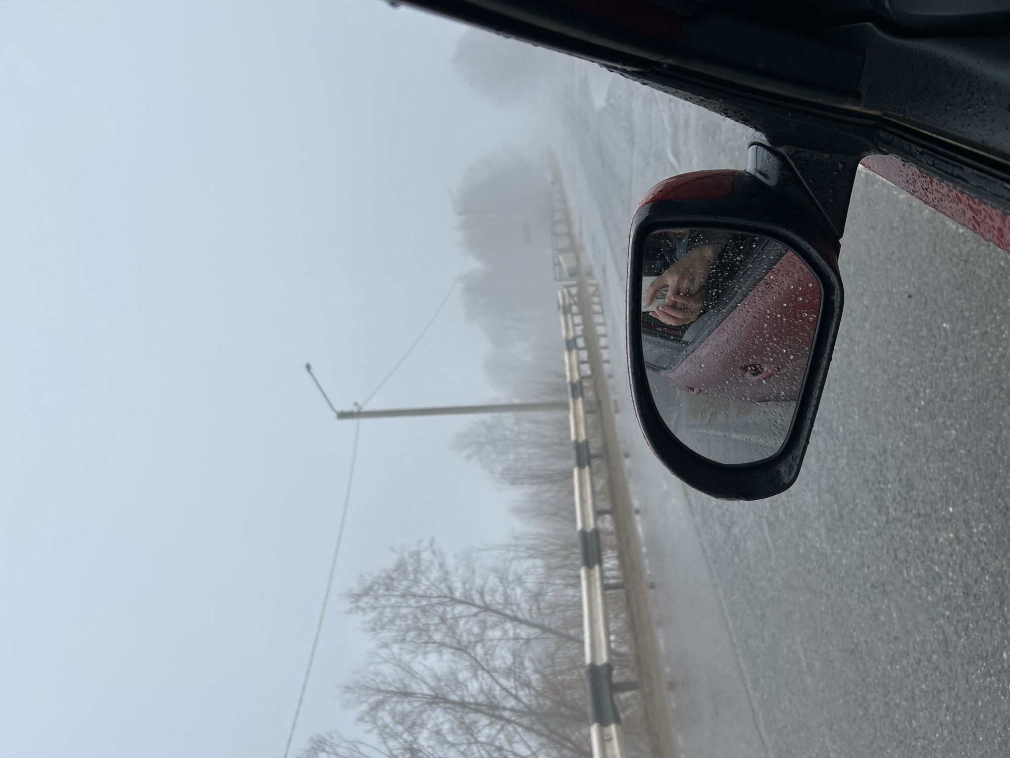 The image shows a rainy, foggy day in Уральское, Russia, viewed from inside a vehicle. The side mirror of a red car is visible, reflecting a person's hands holding a small white object. The road is wet, and there's a metal guardrail. In the distance, there are trees and a street light obscured by fog.