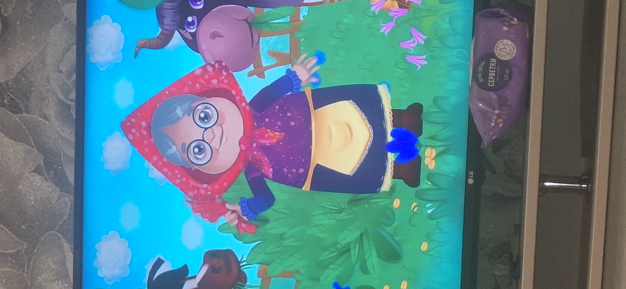 The image shows a television screen displaying a cartoon illustration of a grandmother character wearing a red headscarf and a purple dress with an apron. She is standing in a green, grassy environment with blue sky and white clouds in the background. There are also cartoon animals, including a cow, visible on the screen. Below the television screen, a purple package of wet wipes with Russian text is visible. The television is placed on a stand, and the background behind the television appears to be a textured wall. The image does not contain any information or context related to the city of Bila Tserkva, Ukraine.