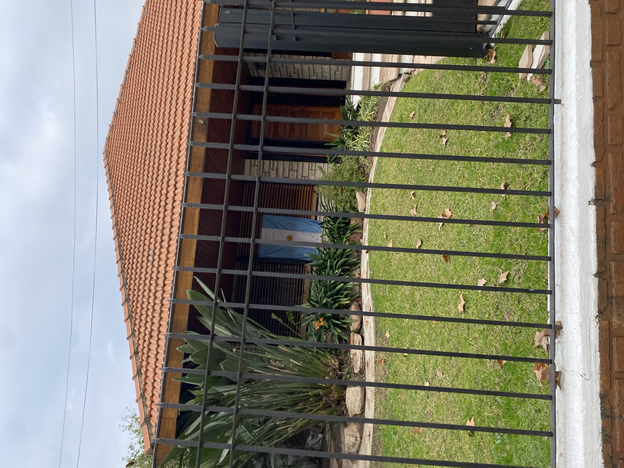 This image shows the front of a house, partially obscured by a black metal fence. The house has a terracotta tiled roof and a stone facade. An Argentinian flag is displayed on the exterior wall of the house. The front yard is covered in green grass, with fallen leaves scattered across it. There are several large plants and bushes in front of the house, and some large rocks placed near the plants. The fence is made of vertical metal bars, with a white-capped brick wall at the bottom. The sky is overcast. The location is identified as Martínez, Argentina, based on the presence of the Argentinian flag.
